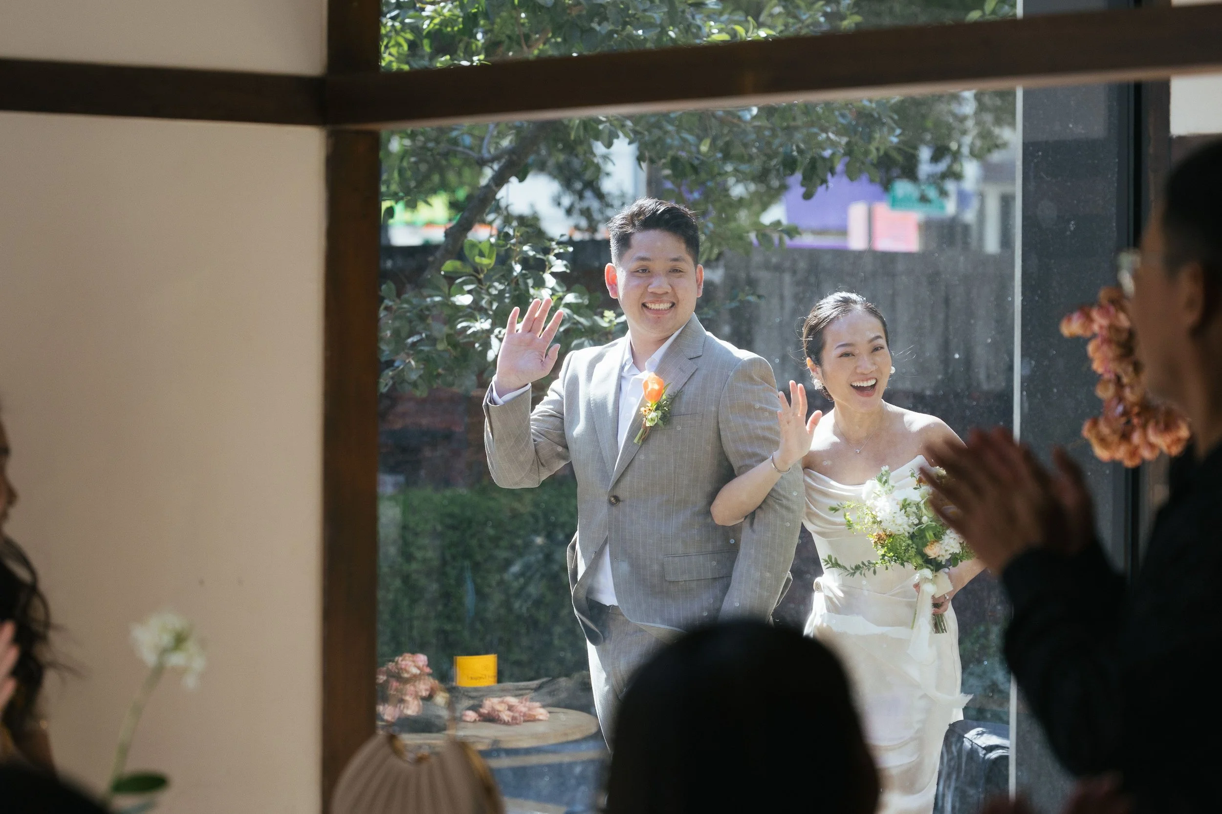 A groom and bride smiling and waving outside a window, greeting guests or friends, during a wedding celebration. The groom is wearing a light gray suit with a boutonnière, and the bride is in a white wedding dress holding a bouquet. They are surround