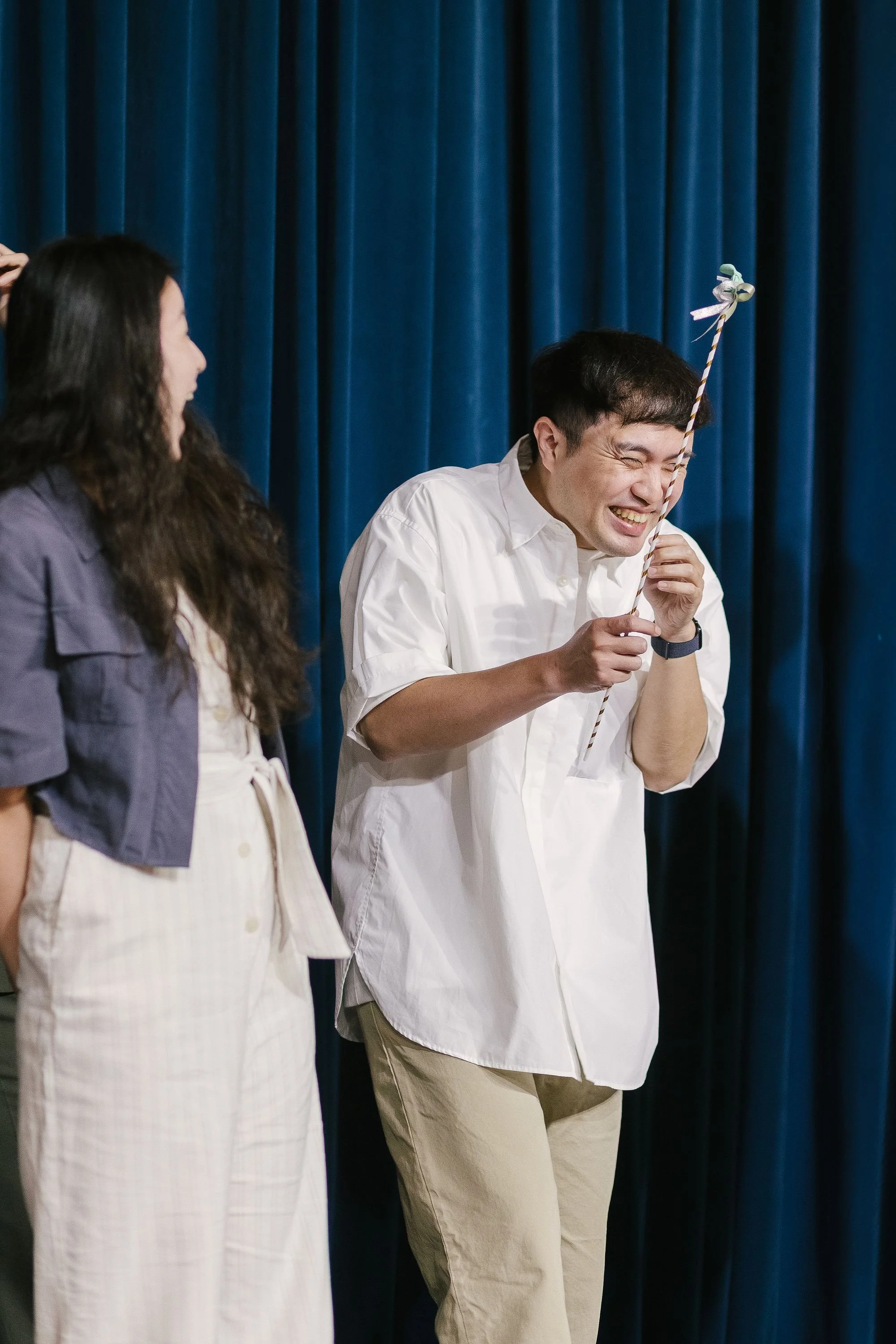 Two people standing in front of a blue curtain, one man holding a decorated stick, both smiling and laughing.