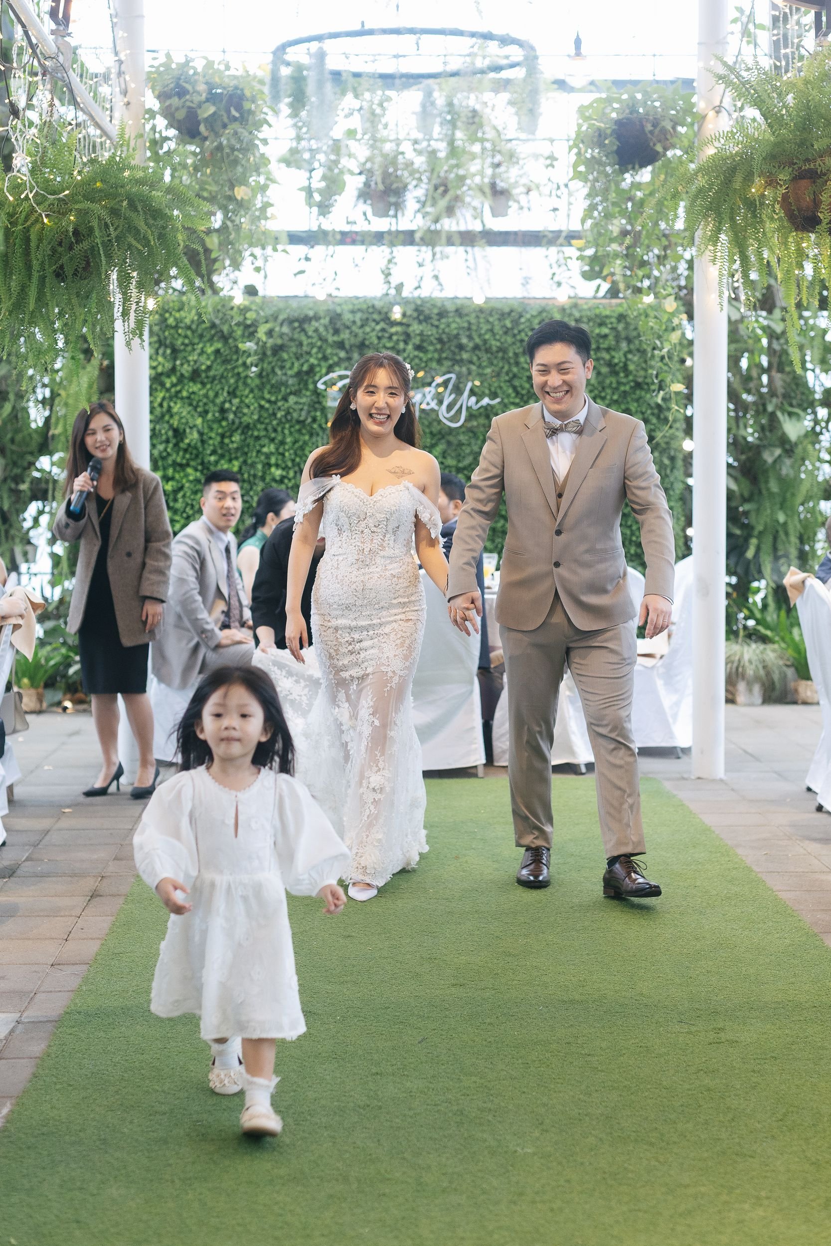 A wedding ceremony with a bride and groom walking on a green aisle, smiling, surrounded by seated guests in a decorated indoor venue with greenery and hanging plants.
