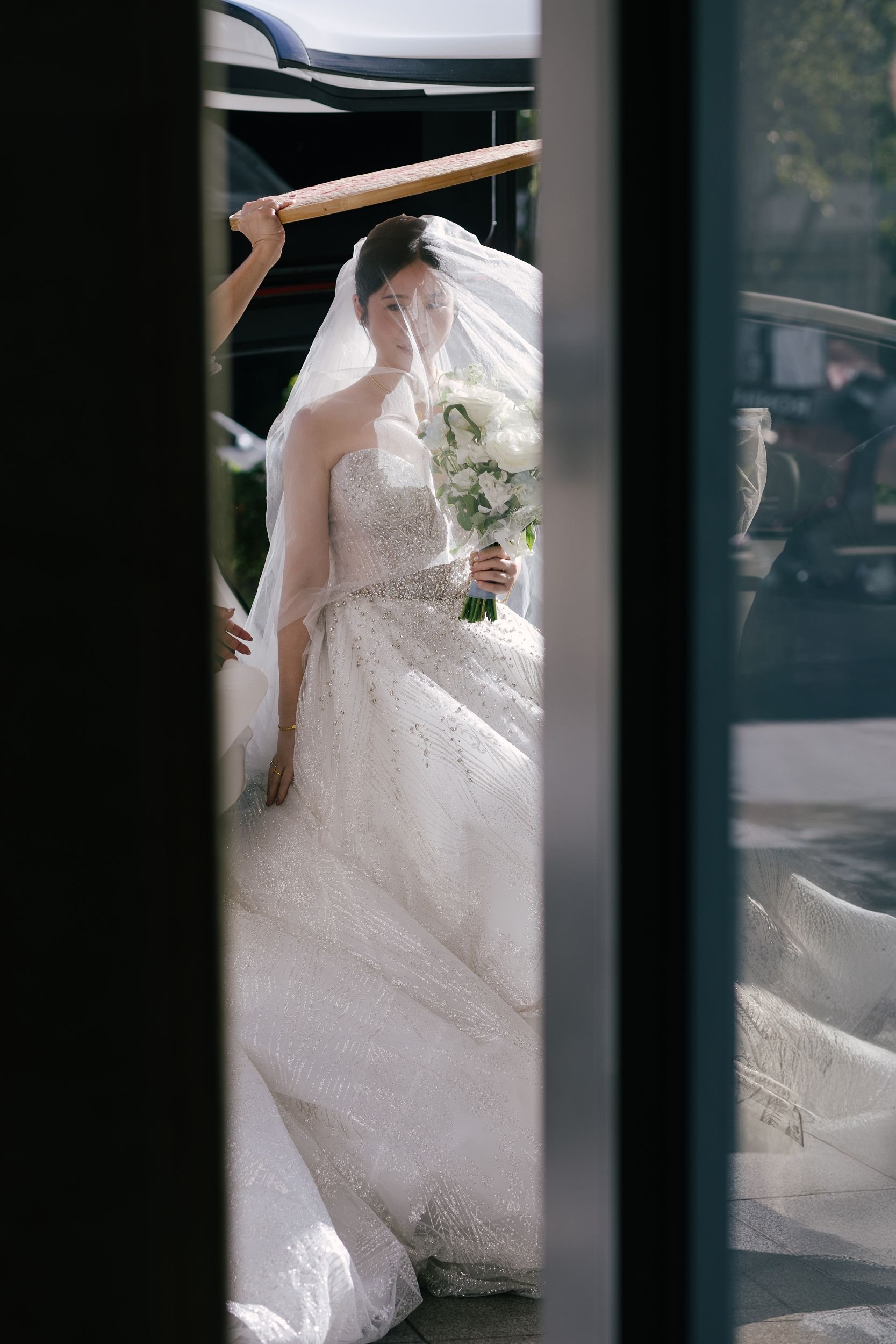 A bride in a white wedding dress with a strapless design and intricate embroidery holds a bouquet of white flowers as she is being helped with her veil by another person. The photo is taken through a narrow gap, possibly a doorway, with sunlight illu