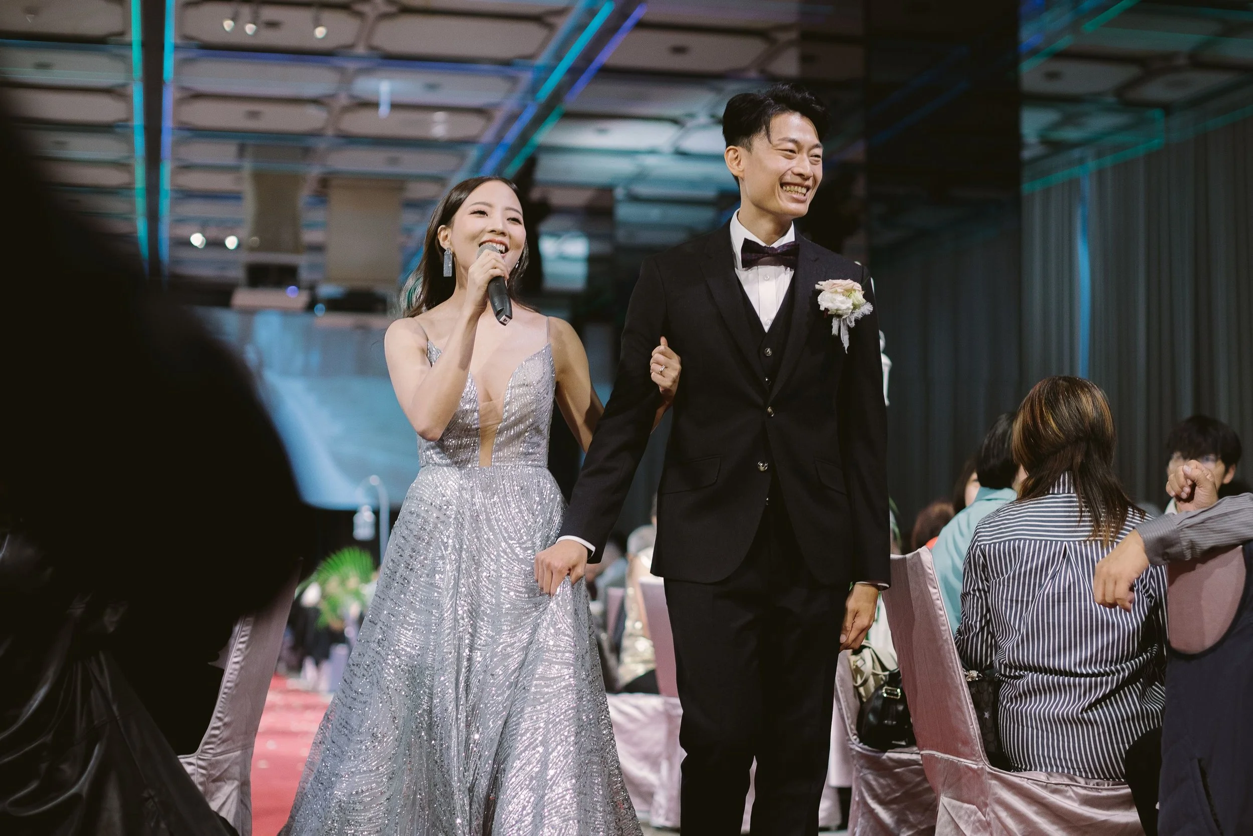A woman in a shimmering silver dress holding a microphone and smiling, standing next to a smiling man in a black tuxedo with a boutonniere, at a formal event or celebration.