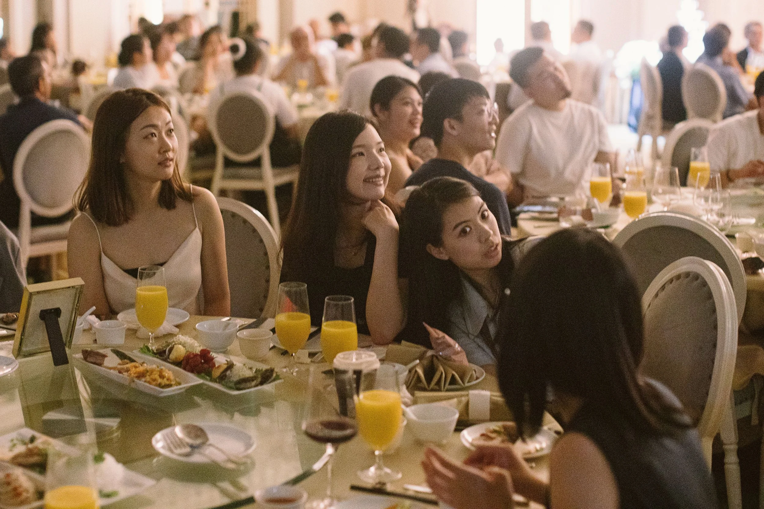 People attending a banquet or dinner event in a large, well-lit room with decorated tables, food, and drinks.