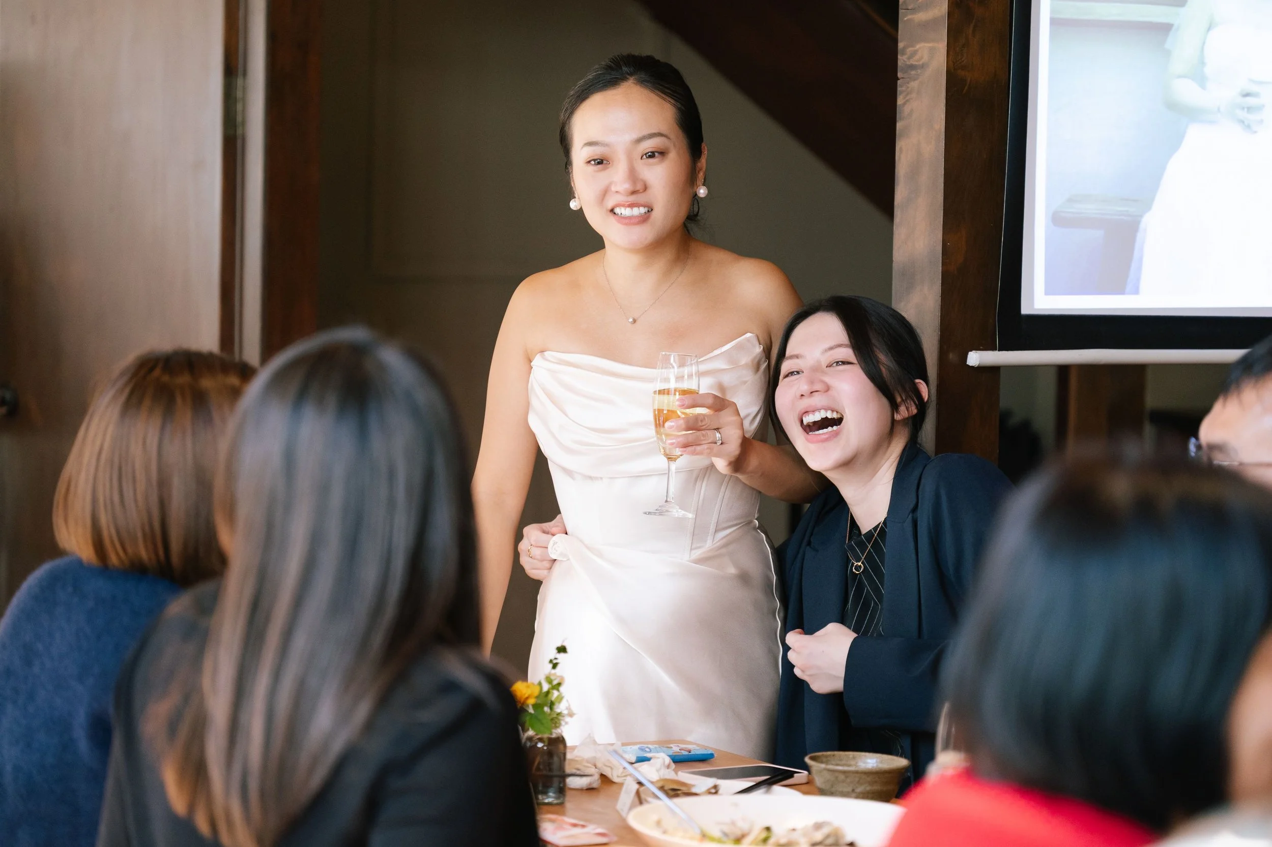 A woman in a white dress holding a glass of champagne stands at a table during a celebration, surrounded by several seated people.