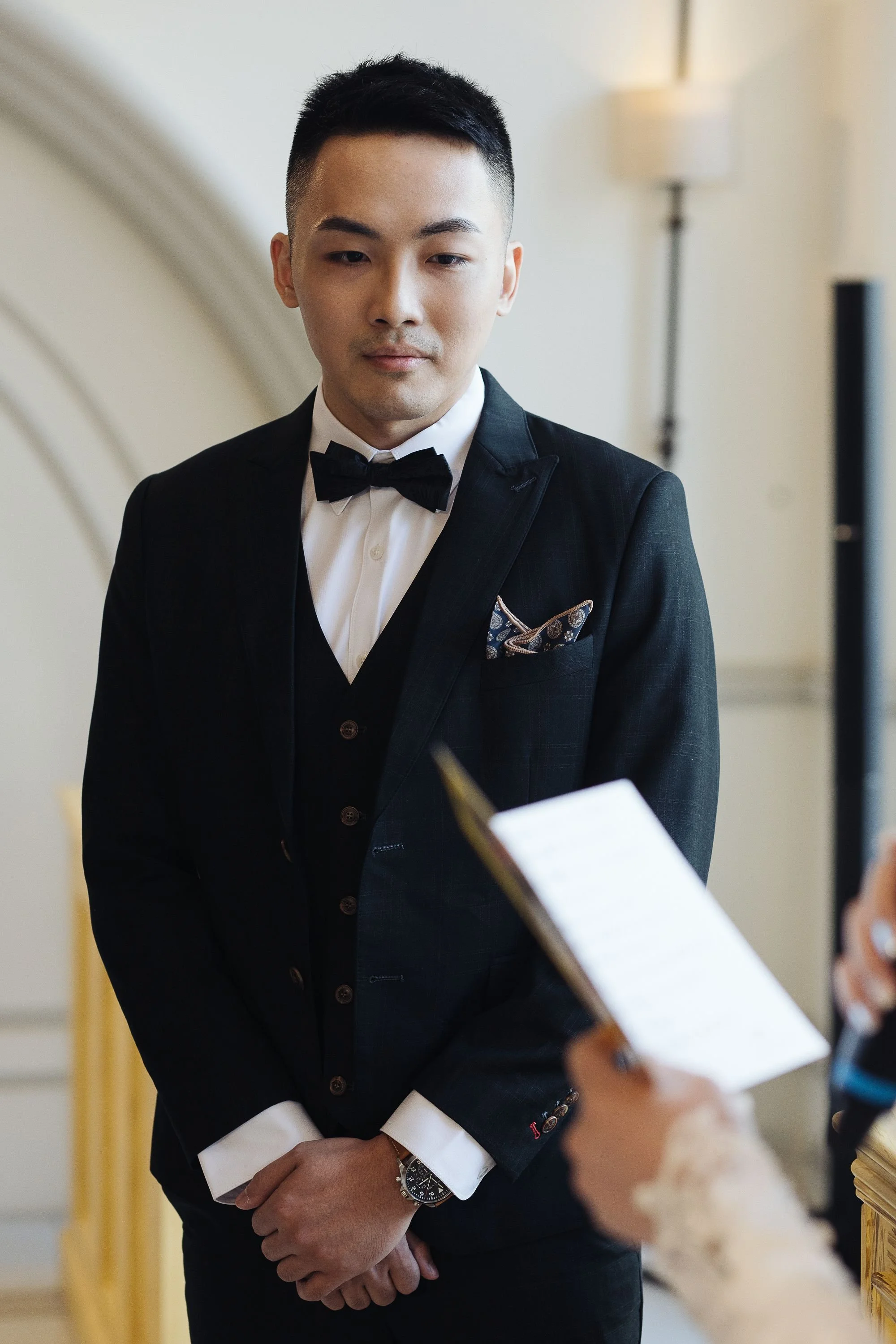 A man dressed in a black tuxedo with a bow tie, standing with his hands clasped, in an indoor setting with cream-colored walls and a standing lamp in the background.