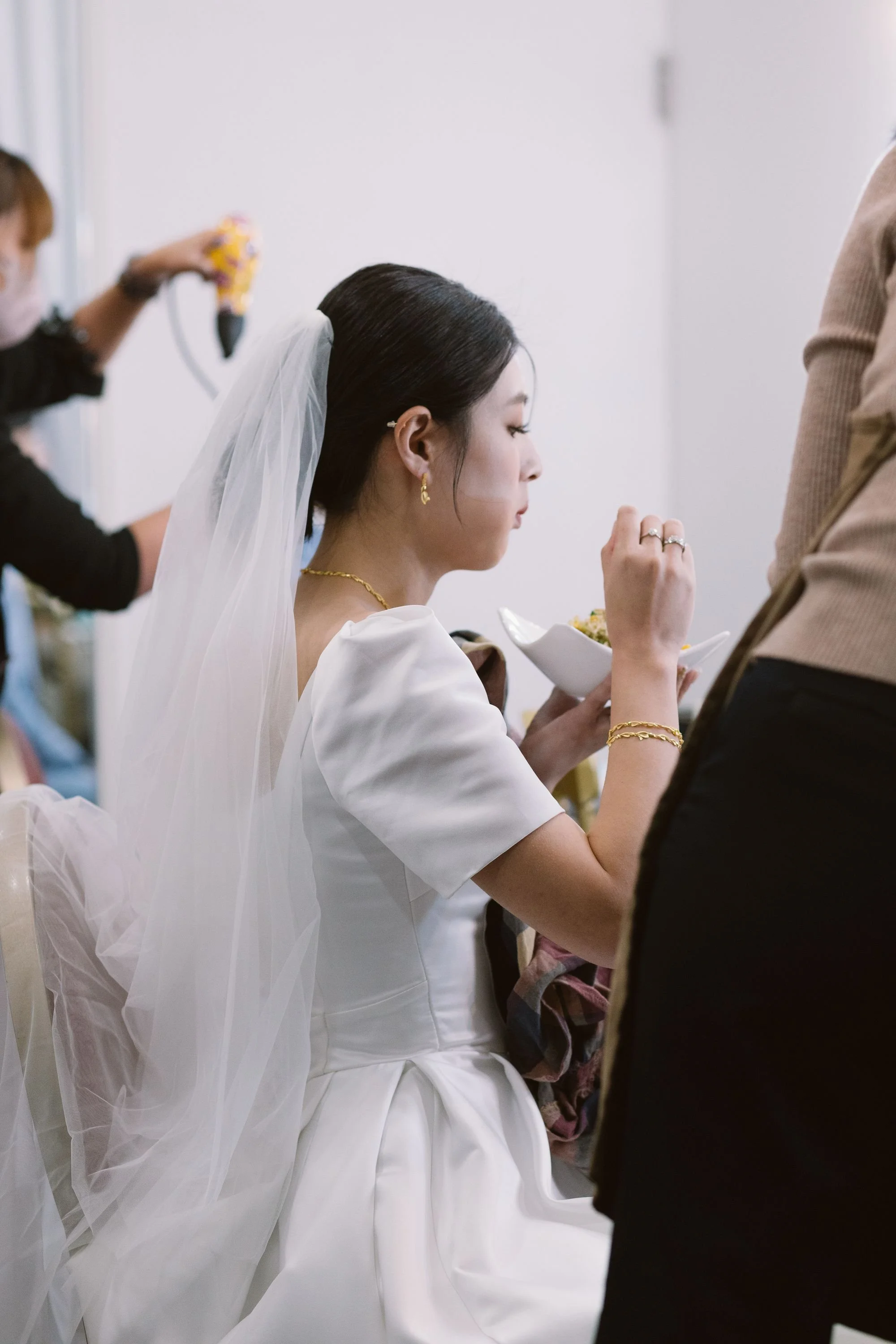 A bride with dark hair styled in an elegant updo, wearing a white wedding dress, gold jewelry, and a veil, is sitting with her eyes closed while eating from a bowl, with a person in the background using a hair styling tool.