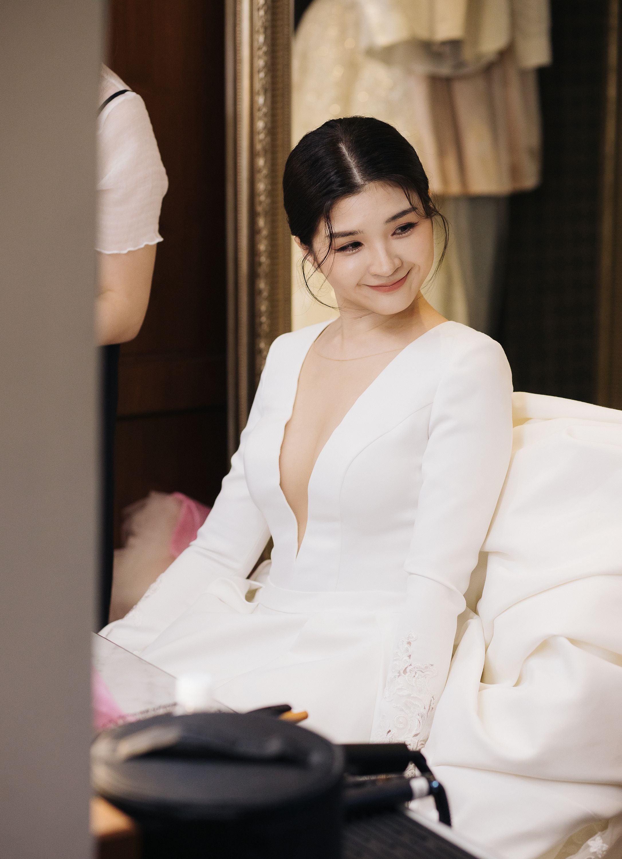 A young woman with dark hair styled in a low bun and wearing a white dress with a plunging neckline, sitting on a cream-colored bed in a well-lit room with gold and dark wood accents, smiling softly and looking away from the camera.