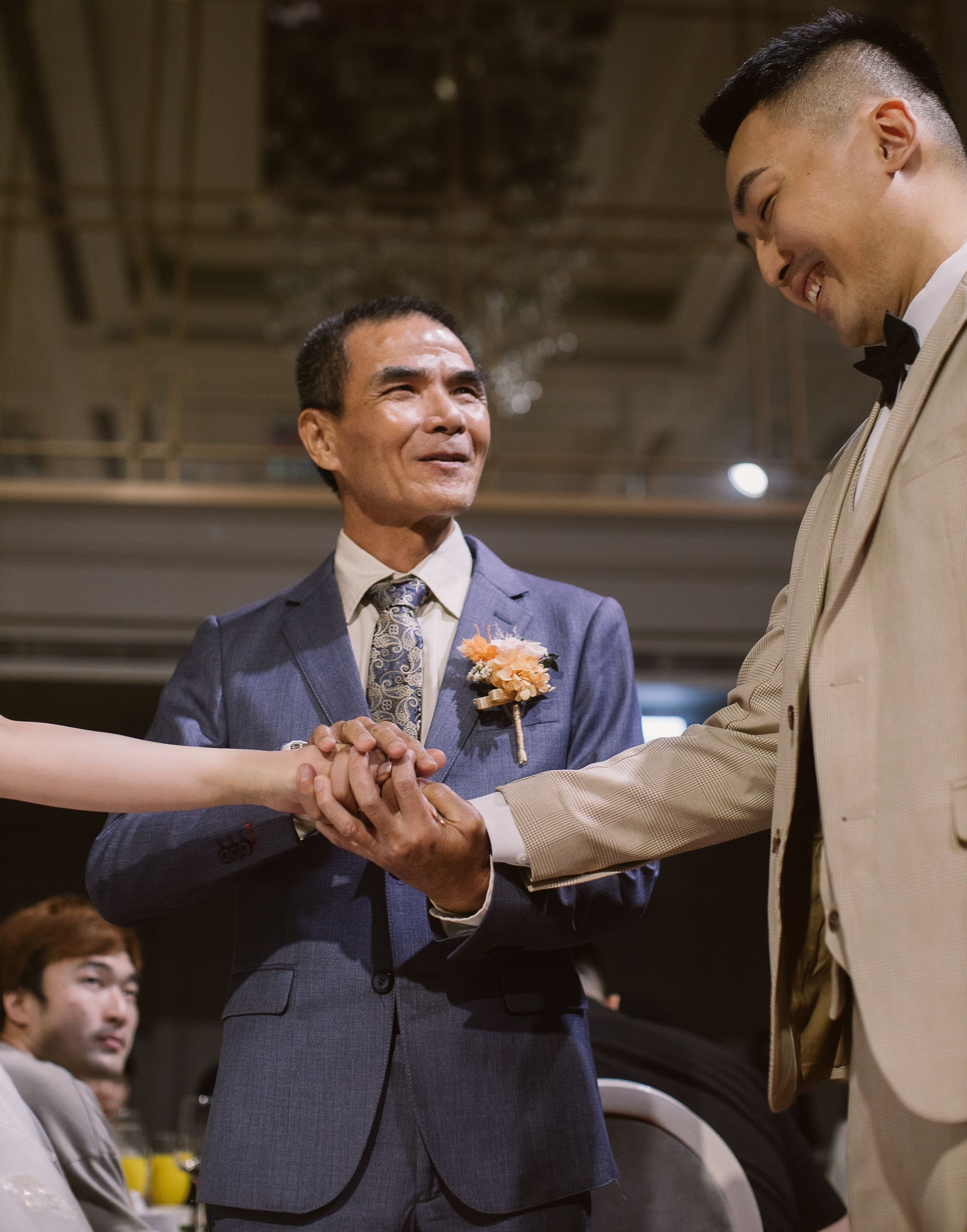 Two men shaking hands at a formal event, with one of them wearing a suit with a boutonniere, and the other in a beige suit with a bow tie, smiling.