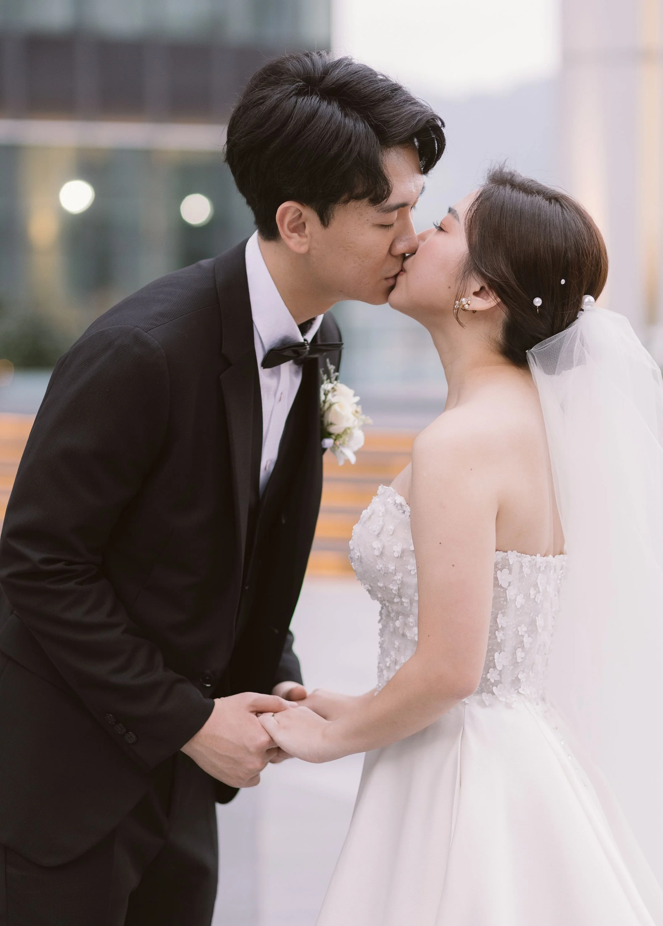 A bride and groom share a kiss while holding hands outdoors.