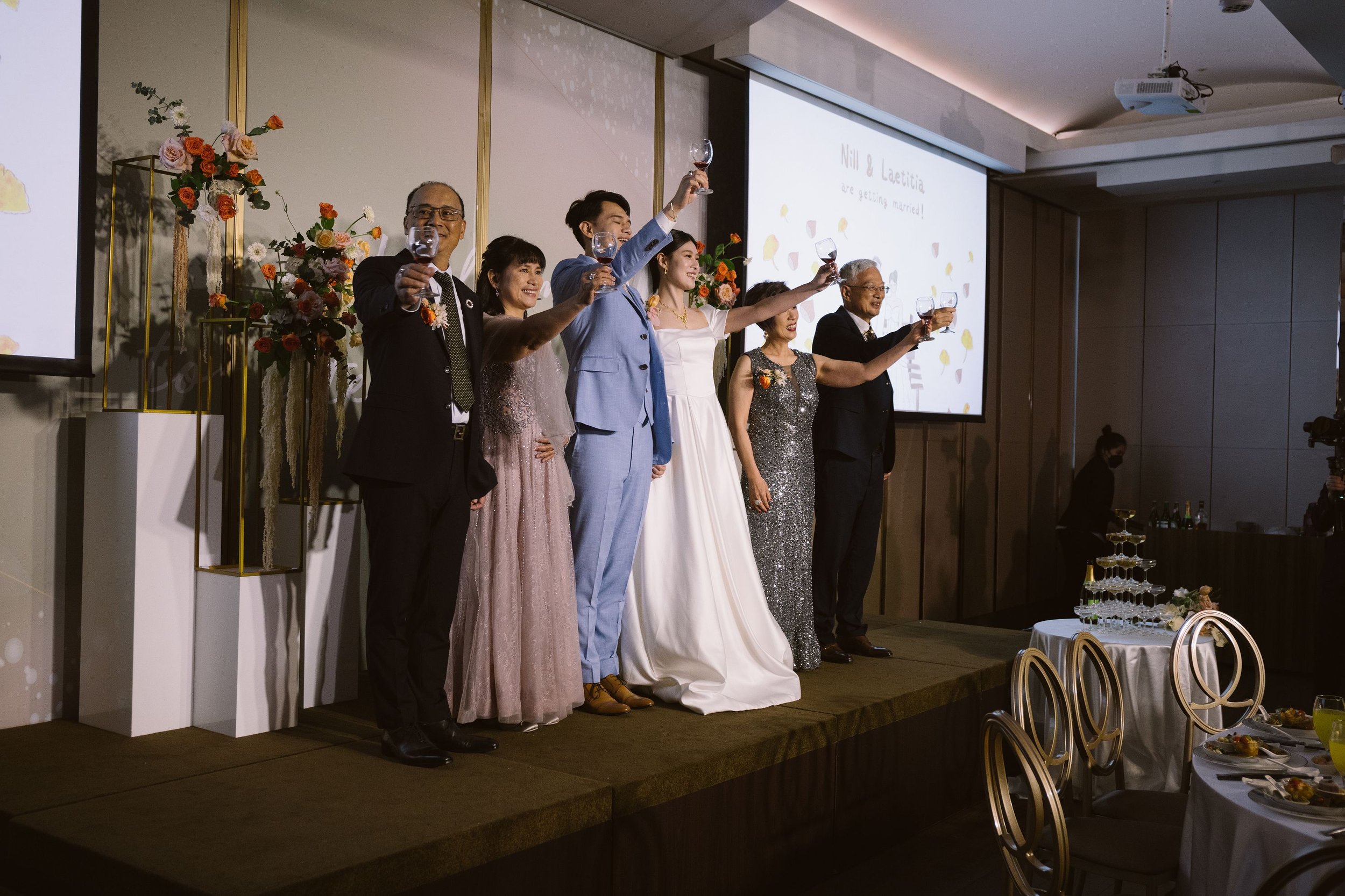 A wedding celebration with a group of people standing on a stage, raising glasses in a toast. The bride is in a white gown, and the groom and guests are dressed in formal attire. There are floral decorations behind them and a large screen displaying 