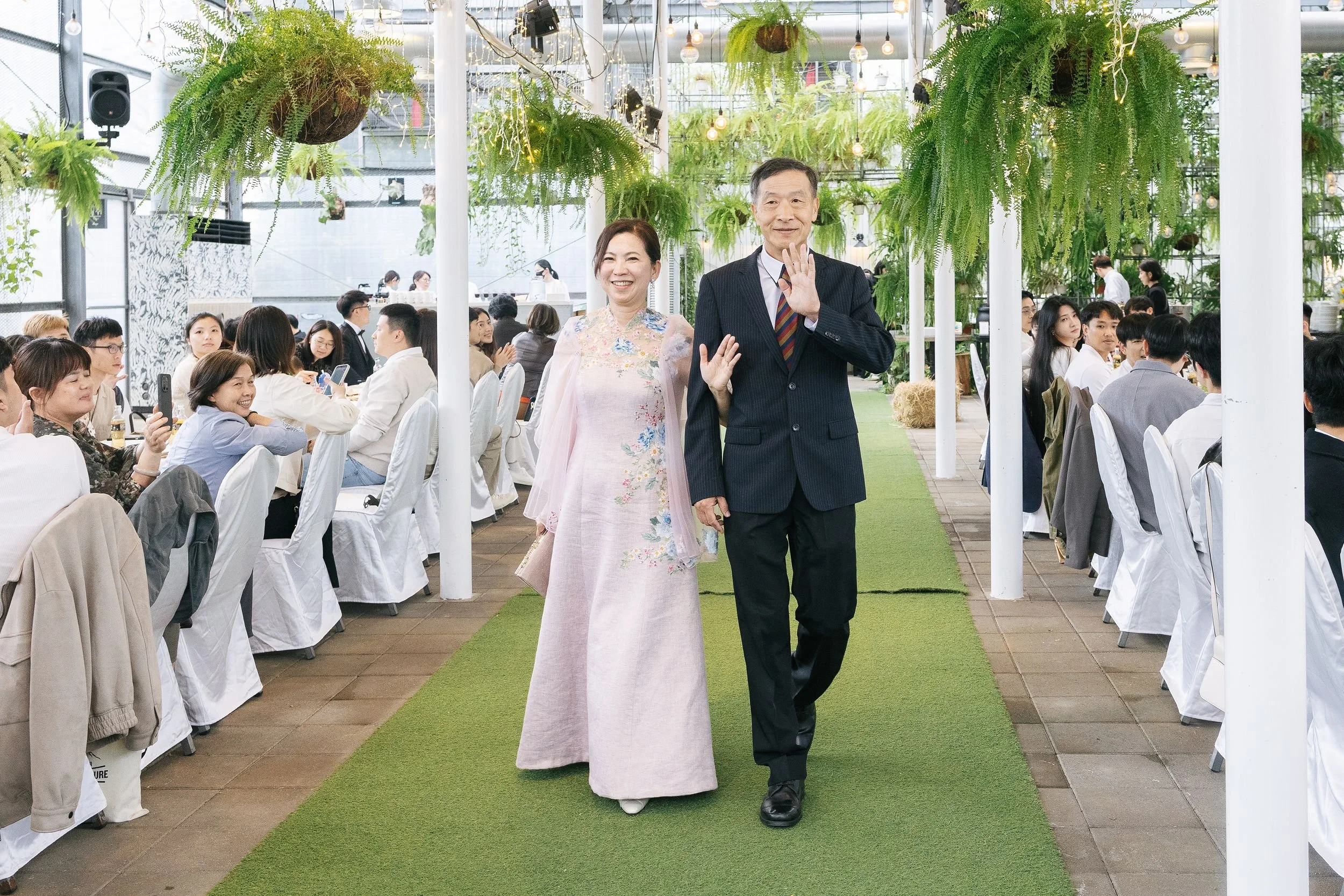 A woman and a man walking down a green carpet at an event, surrounded by seated guests. The woman is wearing a pastel pink floral dress, and the man is in a dark suit with a striped tie. They are smiling and waving, with the woman waving with her rig