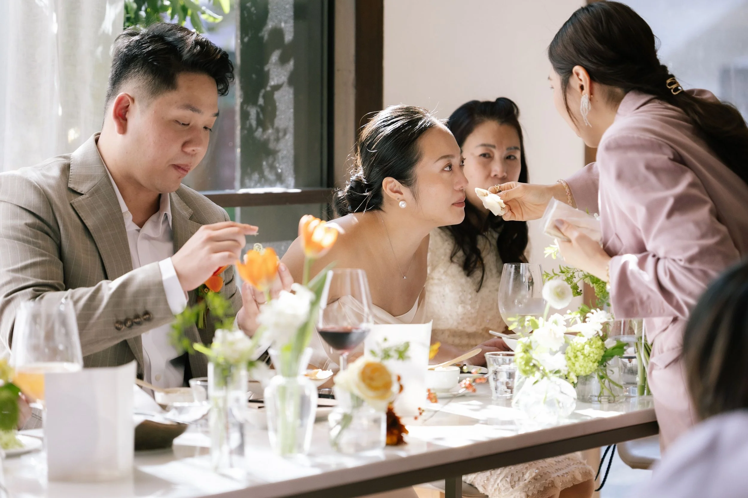 People gathered around a table at a celebration, with a woman in a white dress getting her face painted by a woman in pink. The table has flowers, wine glasses, and food.