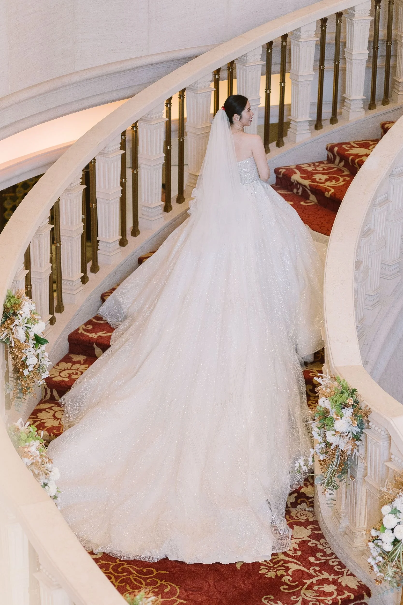 A bride in a white wedding gown and veil standing on a curved staircase with red and gold patterned carpet, floral decorations, and a decorative white railing.