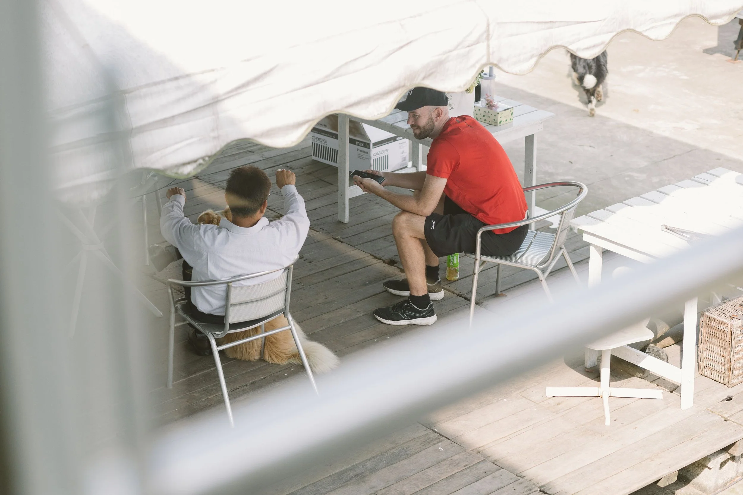A man and a young boy sitting at an outdoor wooden table beneath a tent. The man is wearing a red shirt, black shorts, and a black cap. The boy has dark hair and is wearing a white jacket. There is a dog lying on the ground near them, and a cat is vi