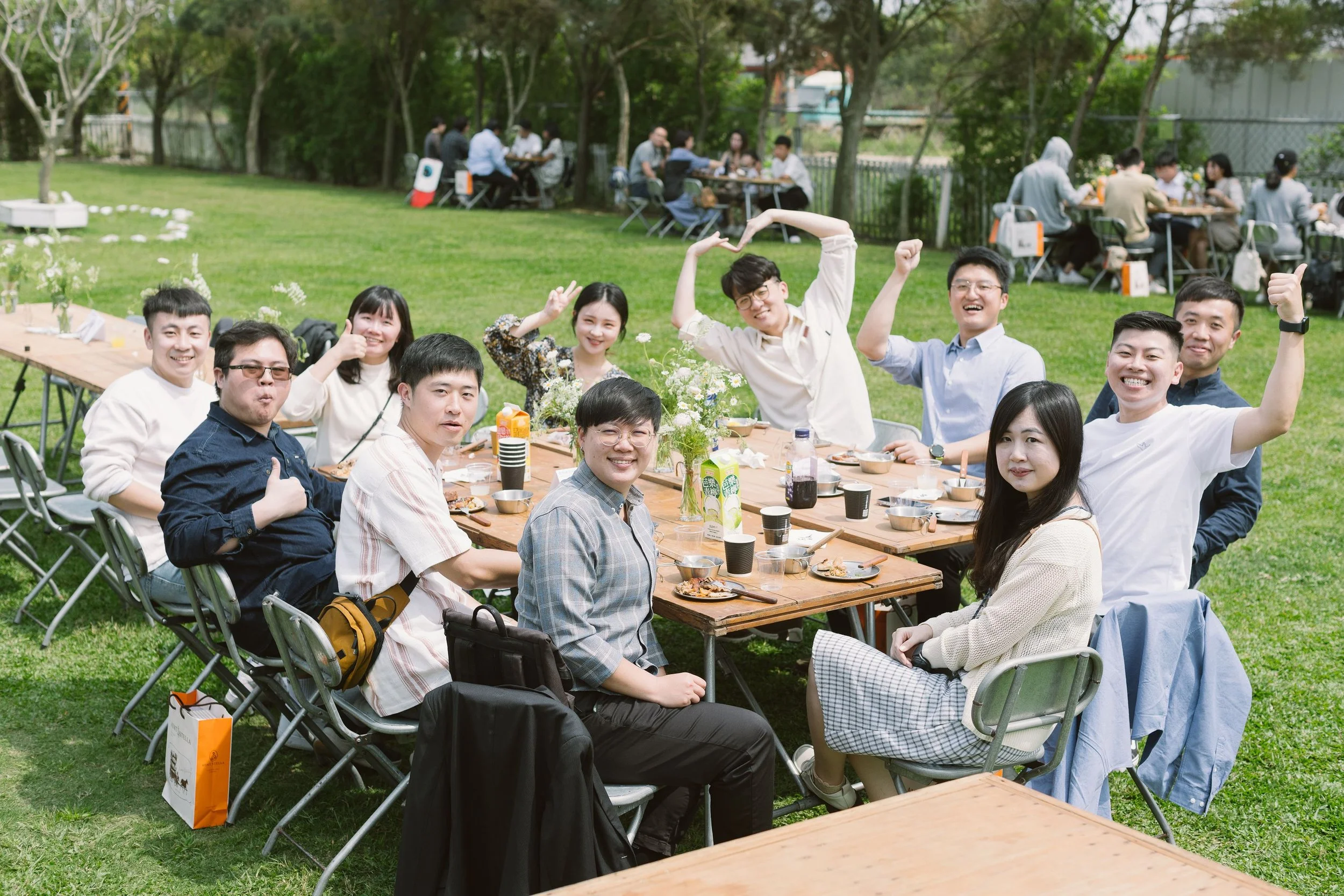 A group of people sitting at a picnic table outdoors, smiling, giving thumbs-up and making heart shapes with their hands, with other groups dining in the background.