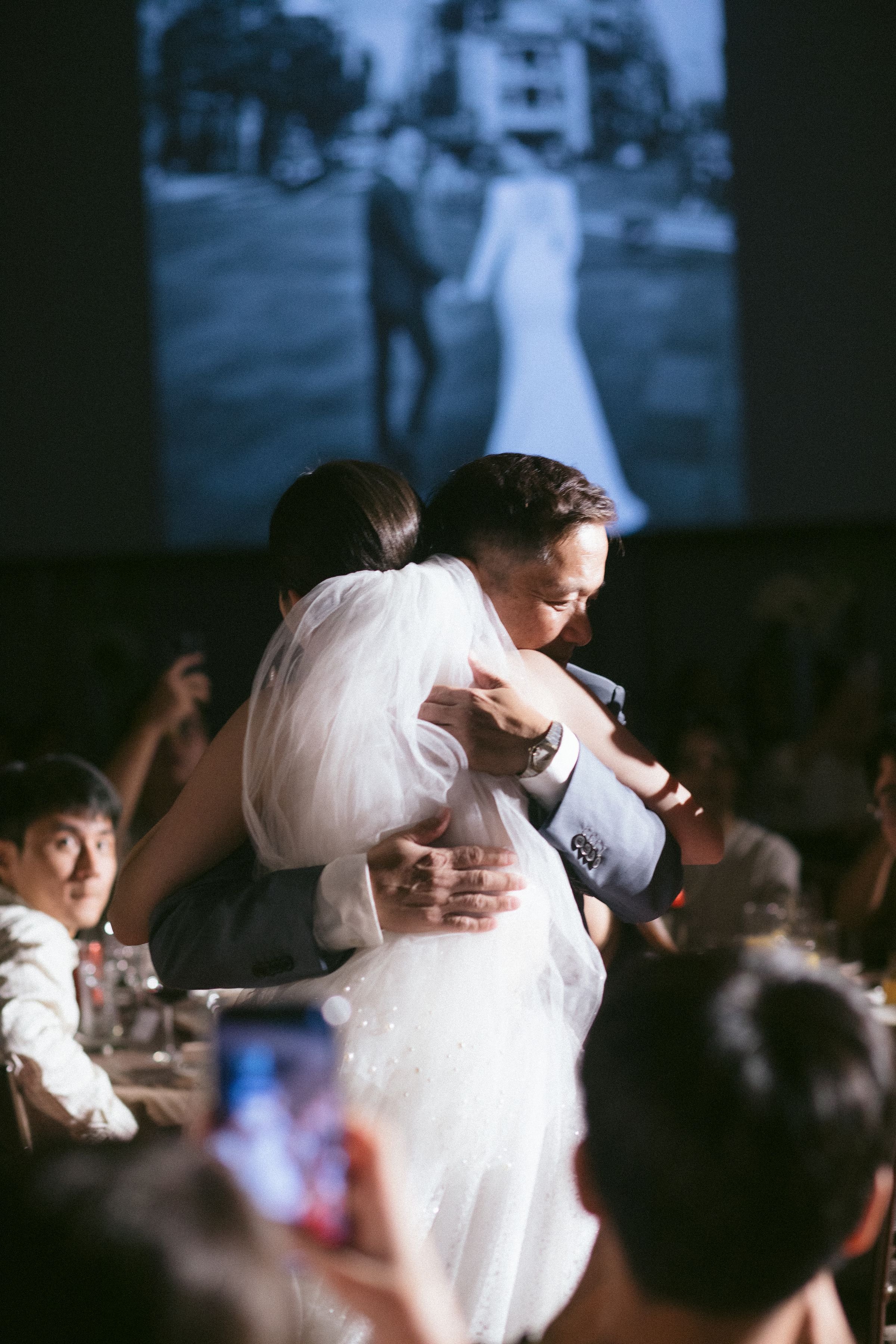 A couple wearing wedding attire sharing a dance at their wedding reception, surrounded by guests, with a large screen showing a blurred image of the couple in the background.
