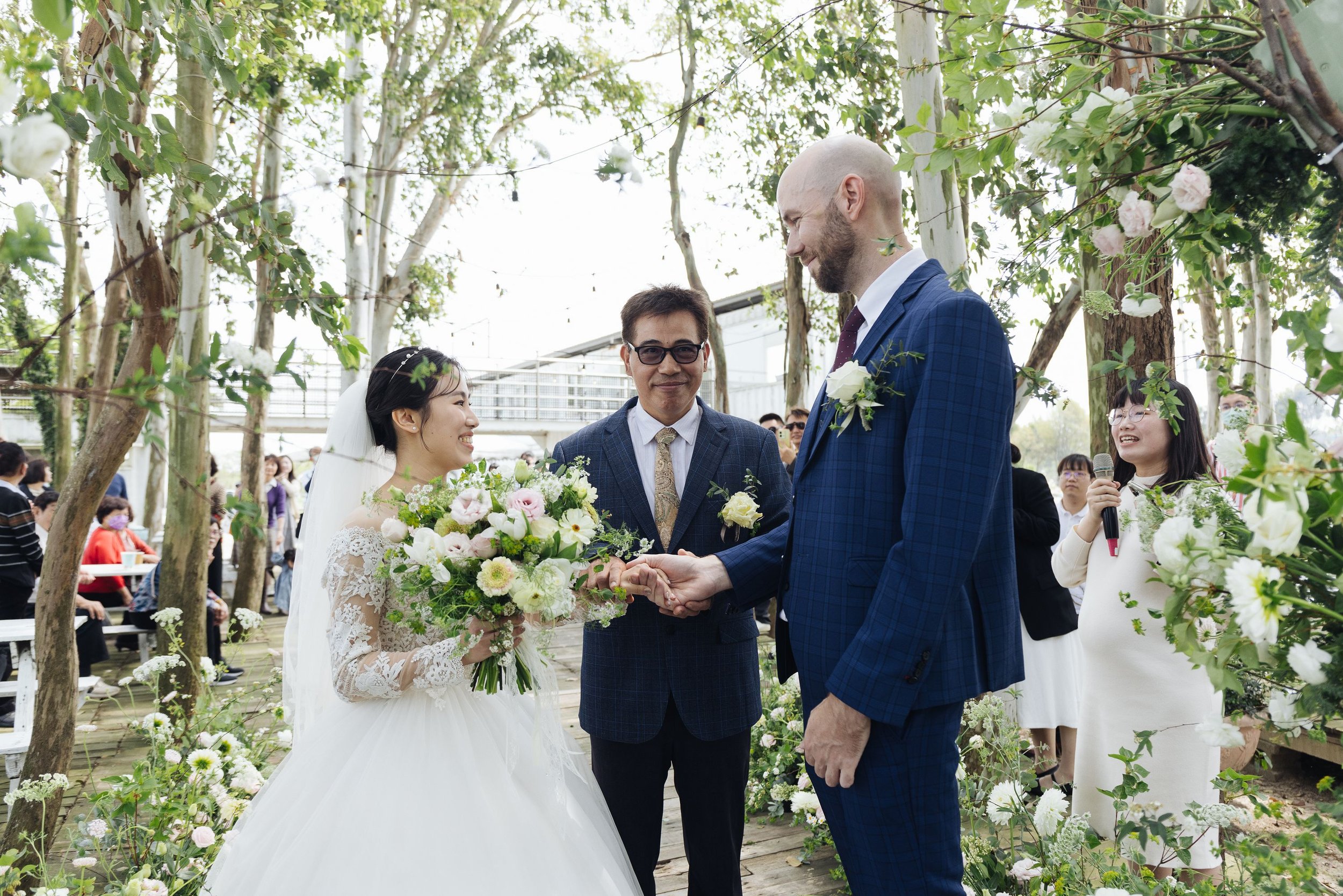 A wedding ceremony outdoors with a bride and groom holding hands, surrounded by floral arrangements and wedding guests. The bride is wearing a lace wedding dress and veil, and the groom is in a blue checkered suit. A woman on the right is holding a m