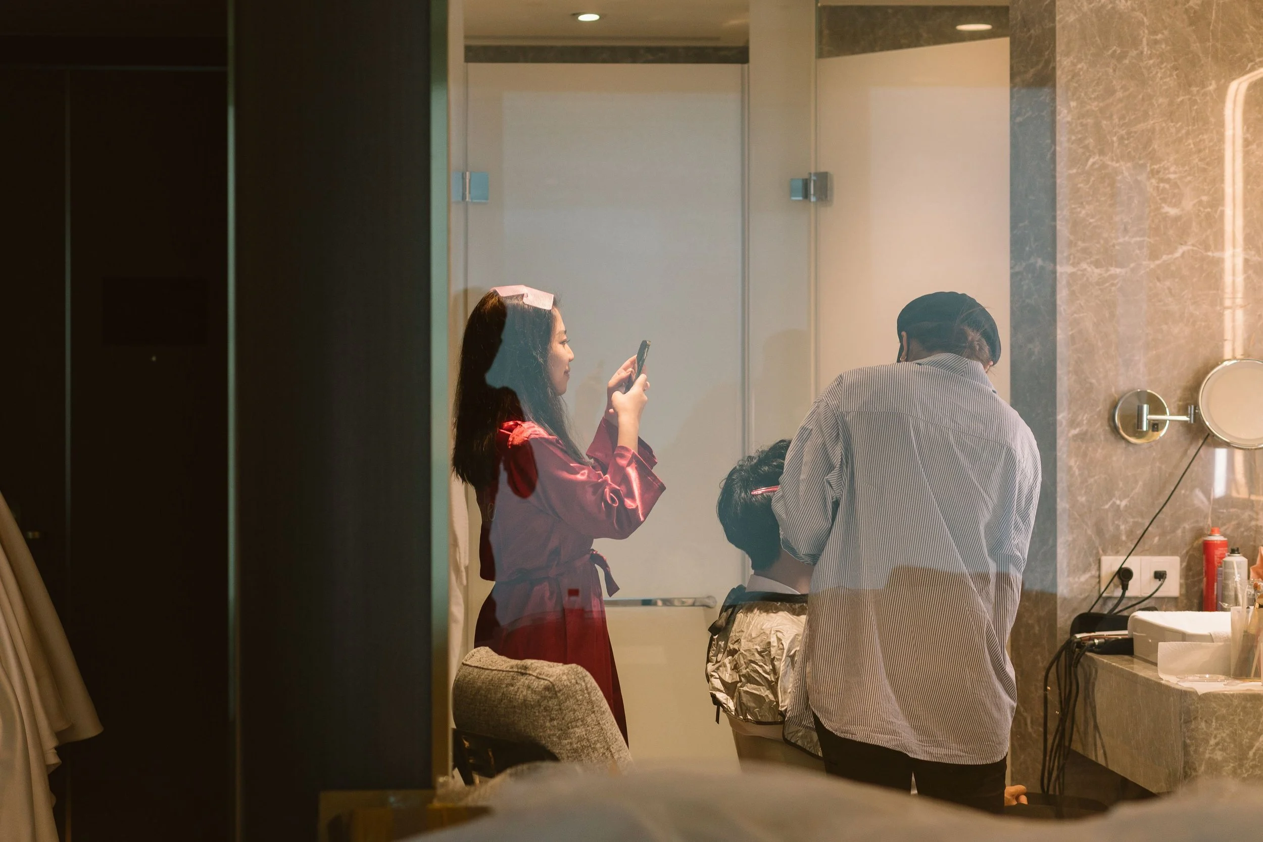 Two women are in a bathroom, one is wearing a pink satin robe with a paper on her head, and the other is wearing a striped shirt. They are looking at a smartphone, and there are various bathroom items on the counter.