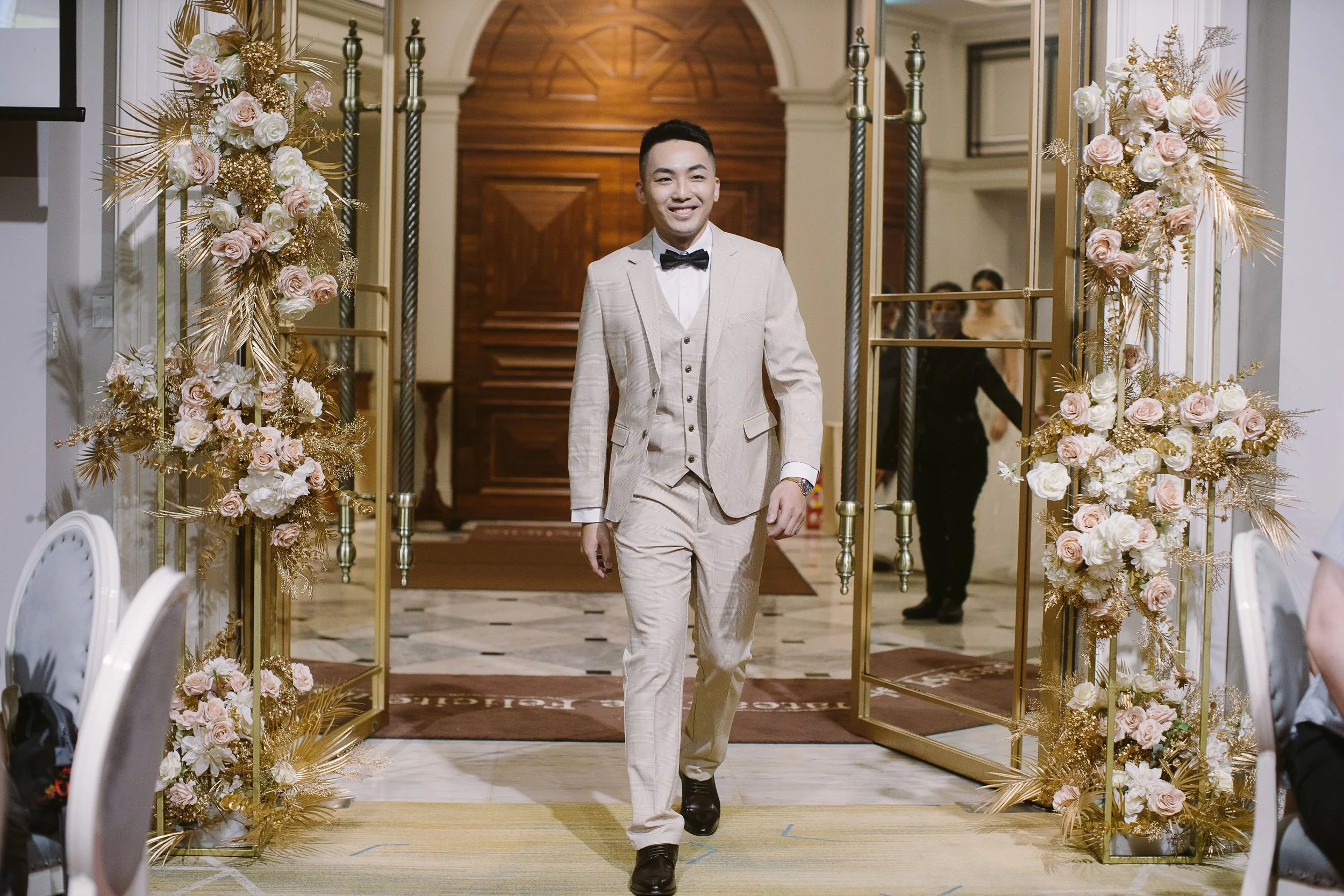 A man in a cream-colored suit with a black bow tie walking through a wedding aisle decorated with pink and white flowers and gold accents, smiling at the camera.