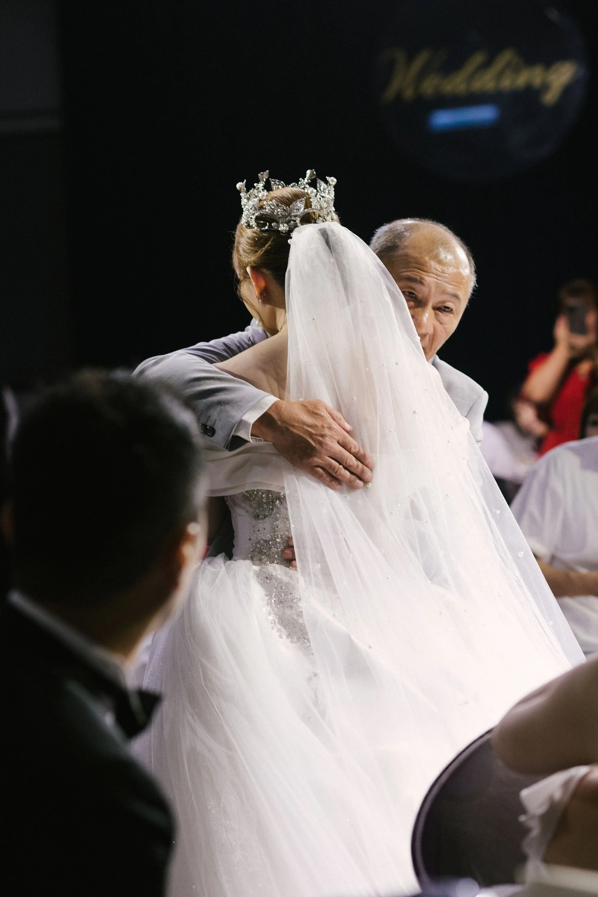 A bride in a wedding gown wearing a tiara and veil being embraced by an older man, possibly her father, at a wedding reception.