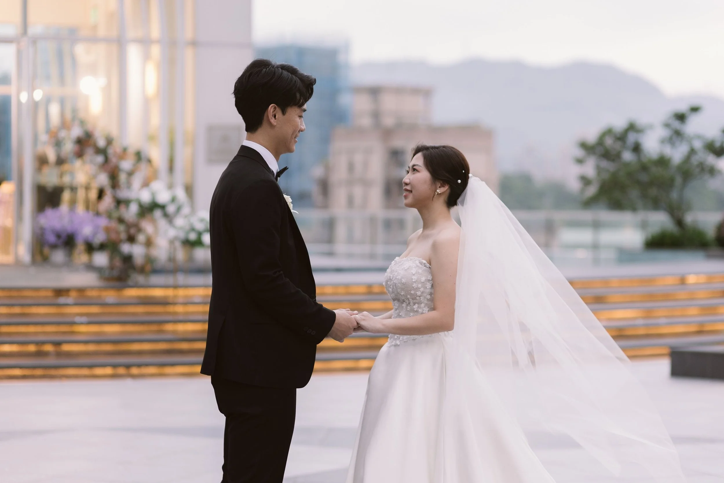 A groom and bride holding hands and smiling at each other outdoors on their wedding day, with city buildings and trees in the background.