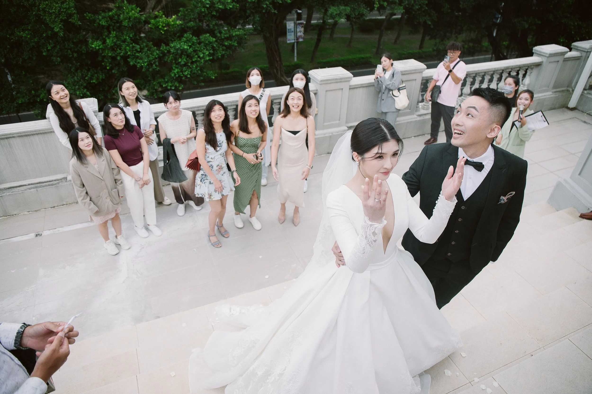 A group of people, including a bride and groom, gathered outdoors for a wedding photo, with some people taking pictures and others smiling.