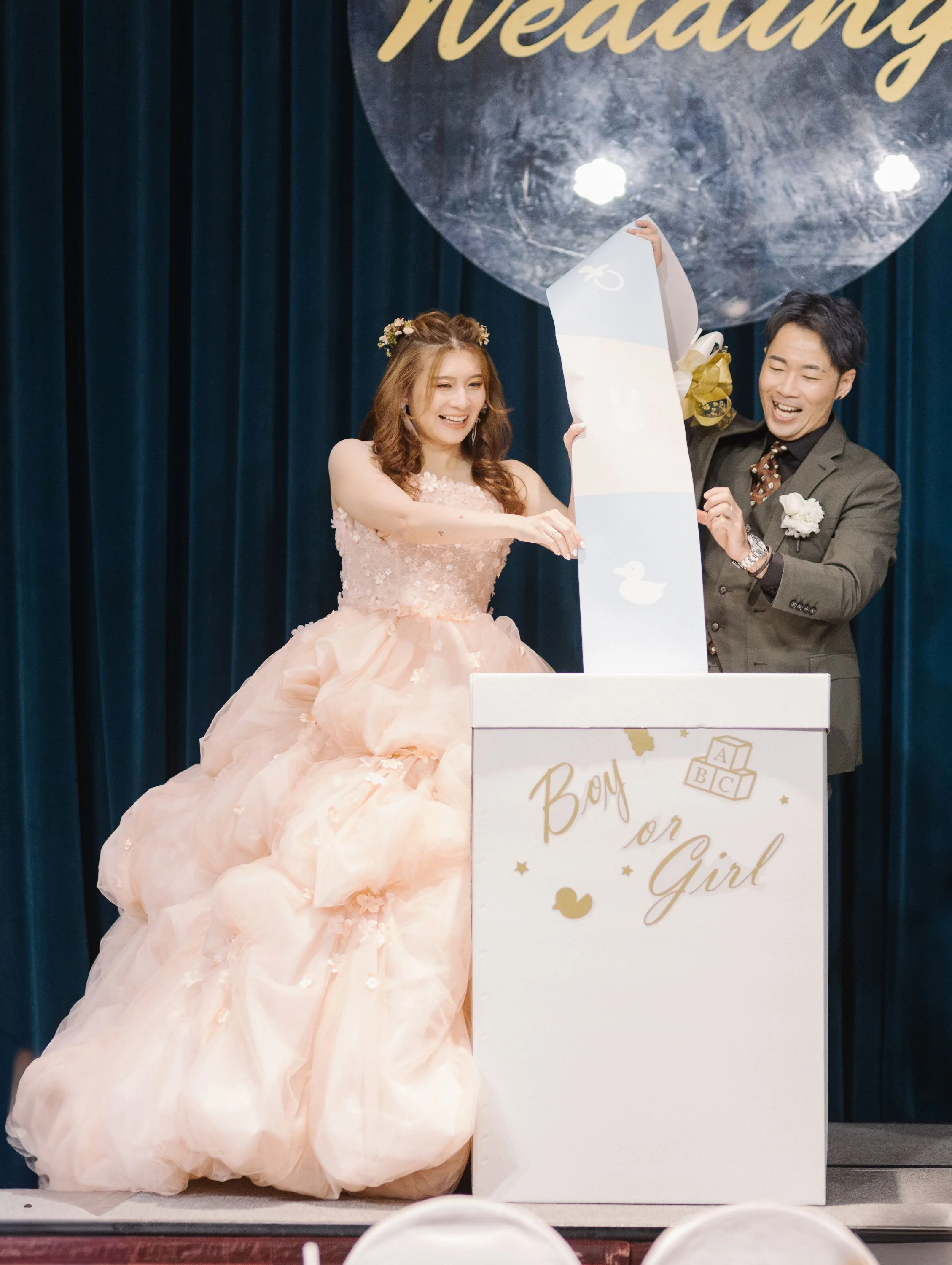 A woman in a pink wedding dress and a man in a suit on stage, smiling and holding a large card with a duck and bunny printed on it. They are at a gender reveal event, with a sign that reads "Boy or Girl" in front of them.