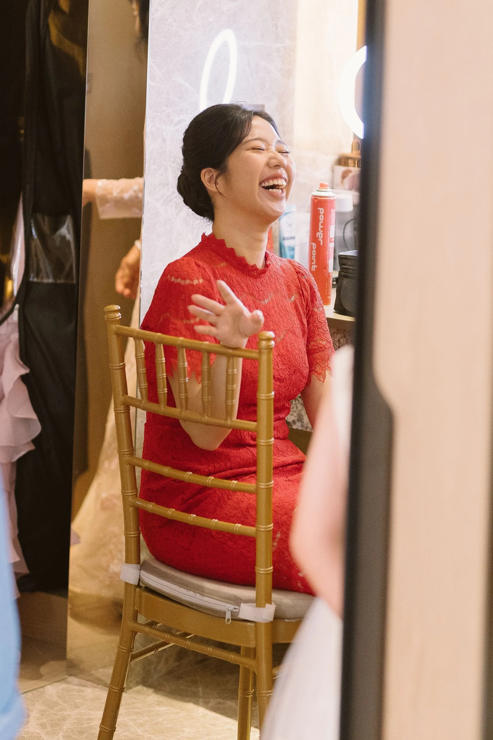A woman with dark hair in an elegant updo, wearing a red lace dress, is smiling and seated on a wooden chair with a rounded back, in front of a mirror in a dressing room or backstage area, with various items visible on a countertop behind her.