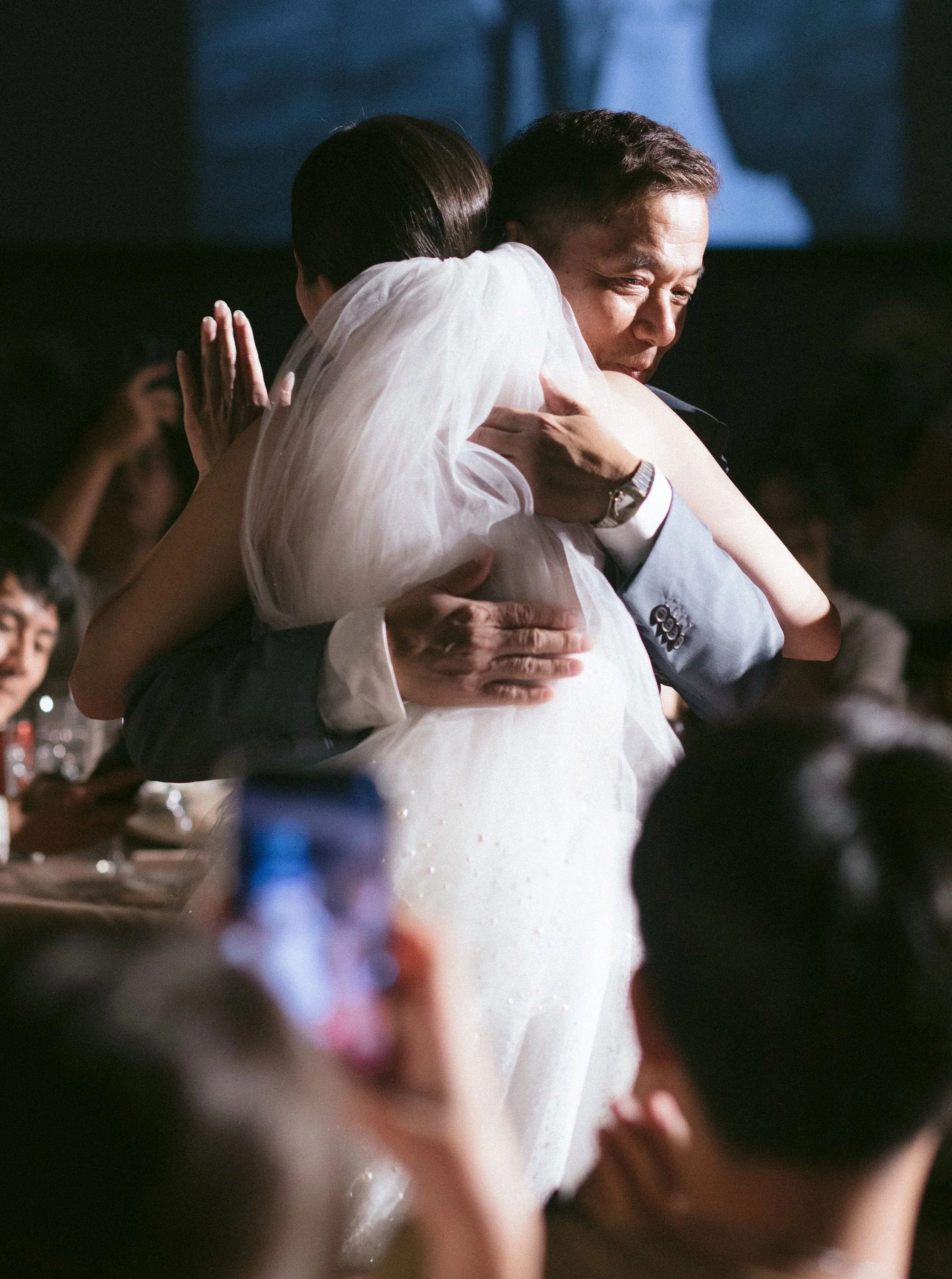 A couple sharing a hug at a wedding reception, with the woman in a white dress and the man in a suit, surrounded by guests.