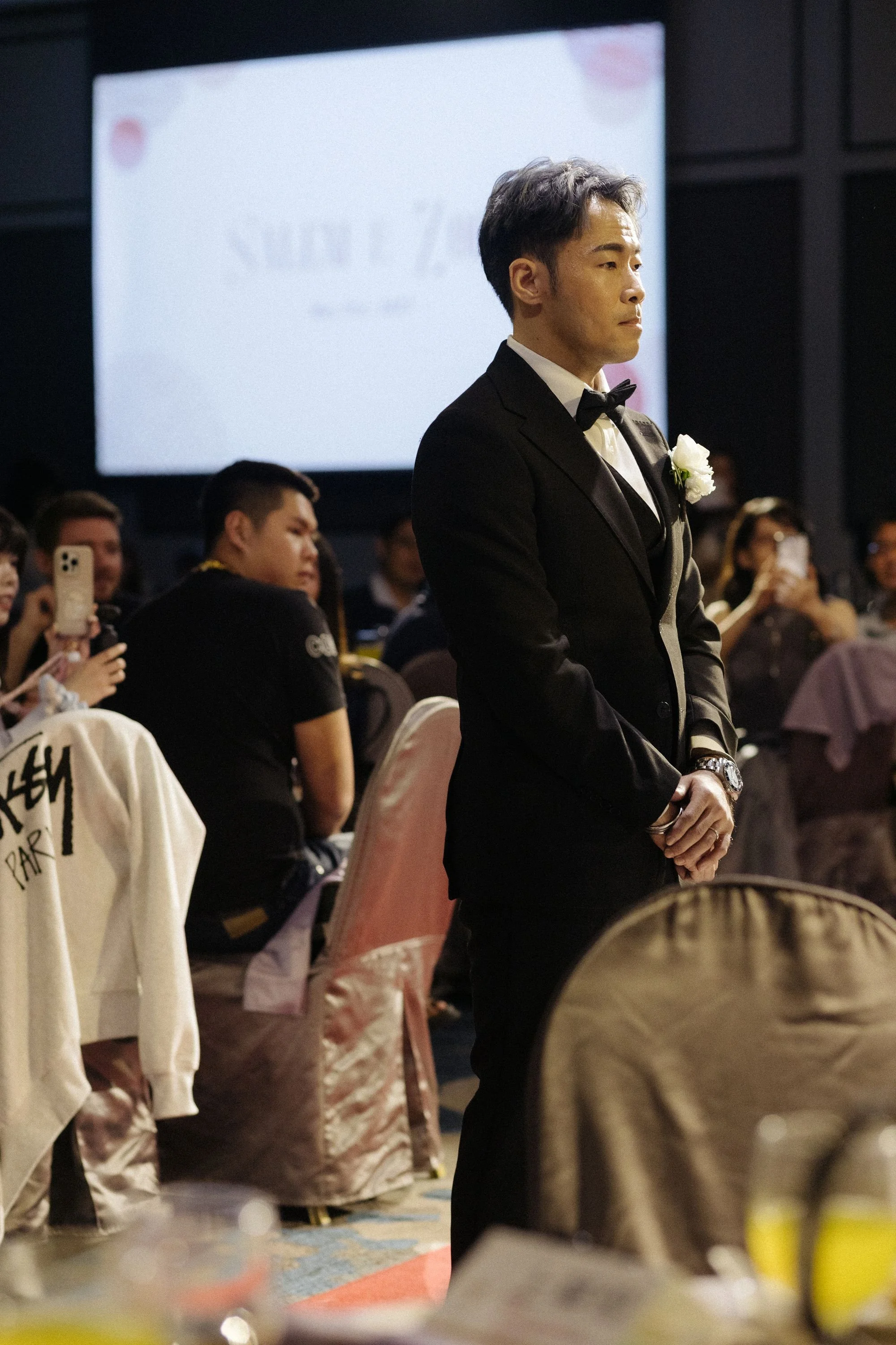 A young man in a tuxedo and bow tie with a white flower on his lapel, standing in profile during a formal event, with guests and a large screen in the background.