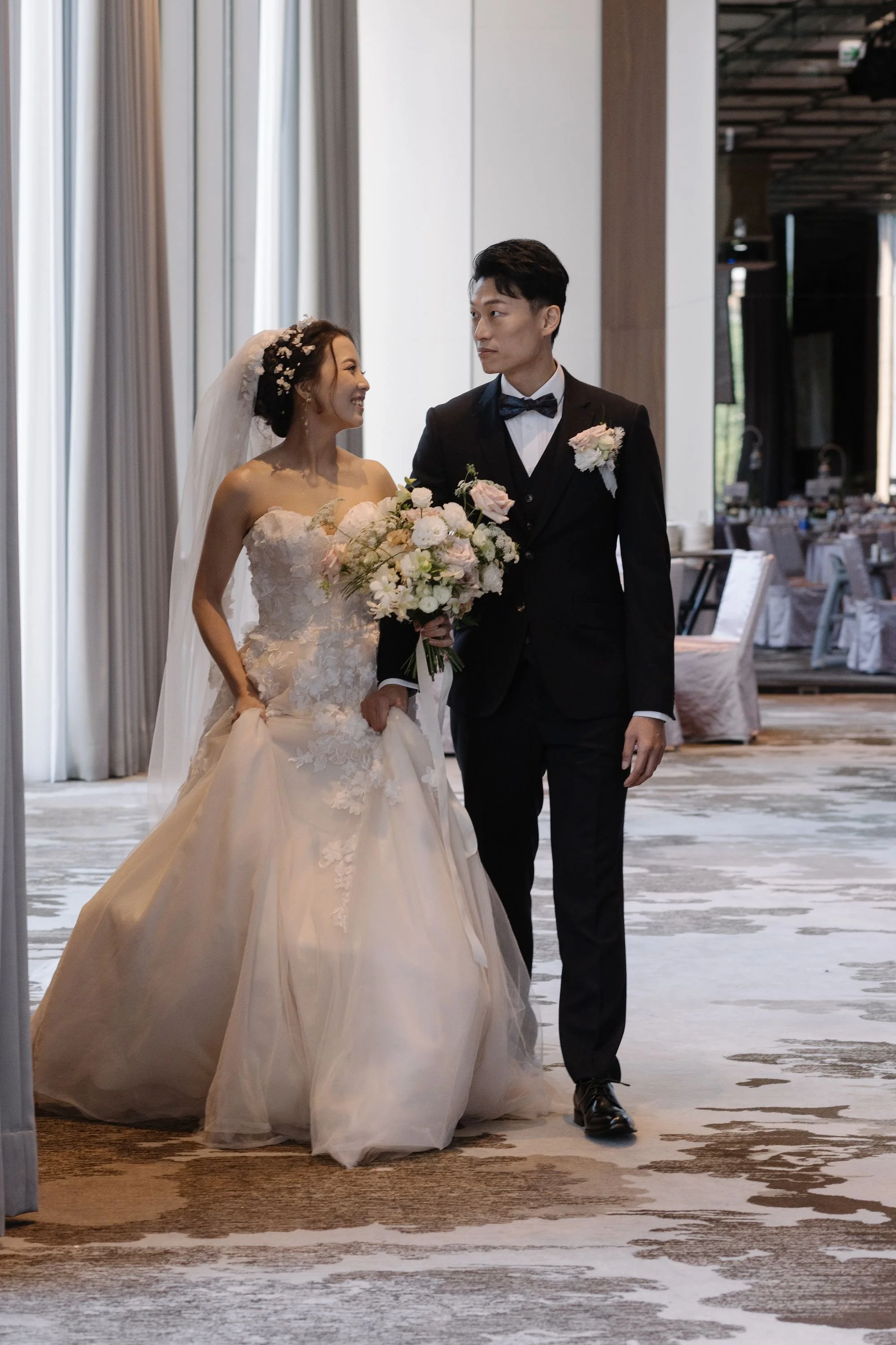 A bride and groom walking together indoors during their wedding reception. The bride is wearing a strapless wedding gown with floral lace details and holding a bouquet of flowers. The groom is dressed in a formal black tuxedo with a bow tie and bouto