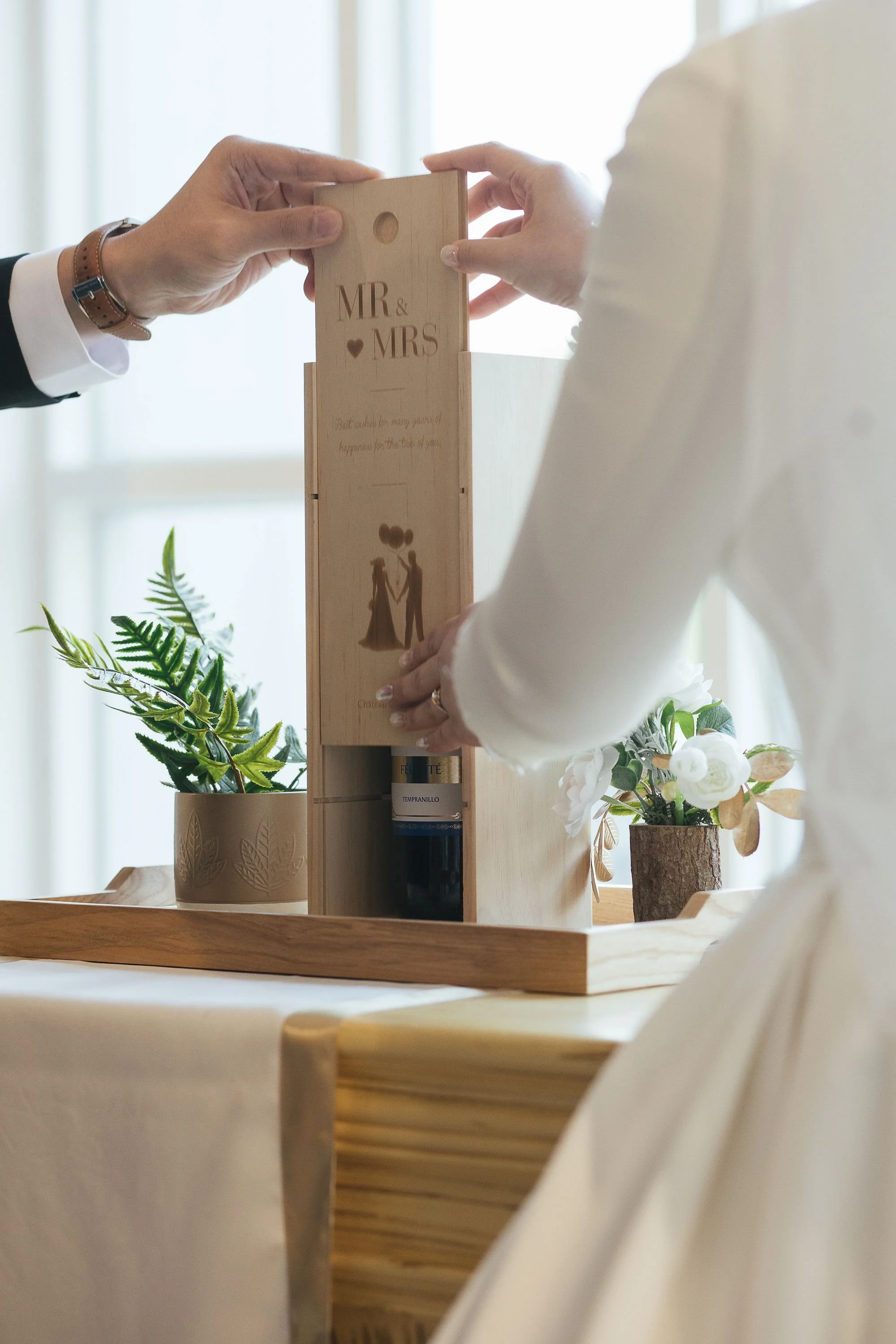 A person placing a wooden 'Mr & Mrs' sign on a wine rack, surrounded by decorative plants on a wooden table.