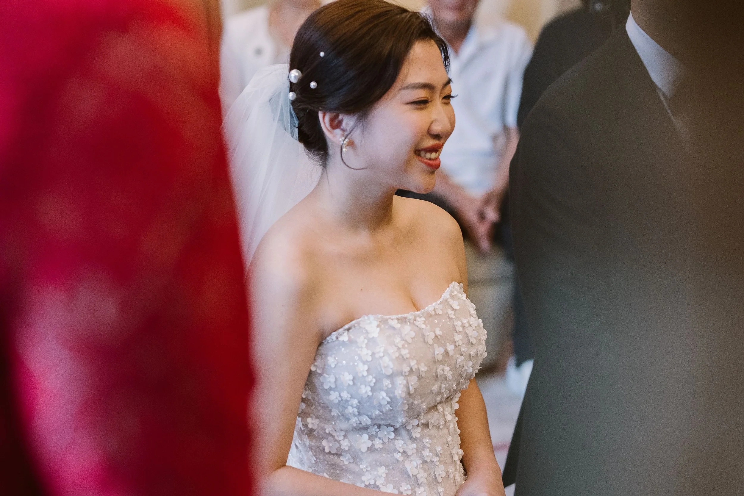 A smiling young woman in a white, floral wedding dress during a wedding ceremony.
