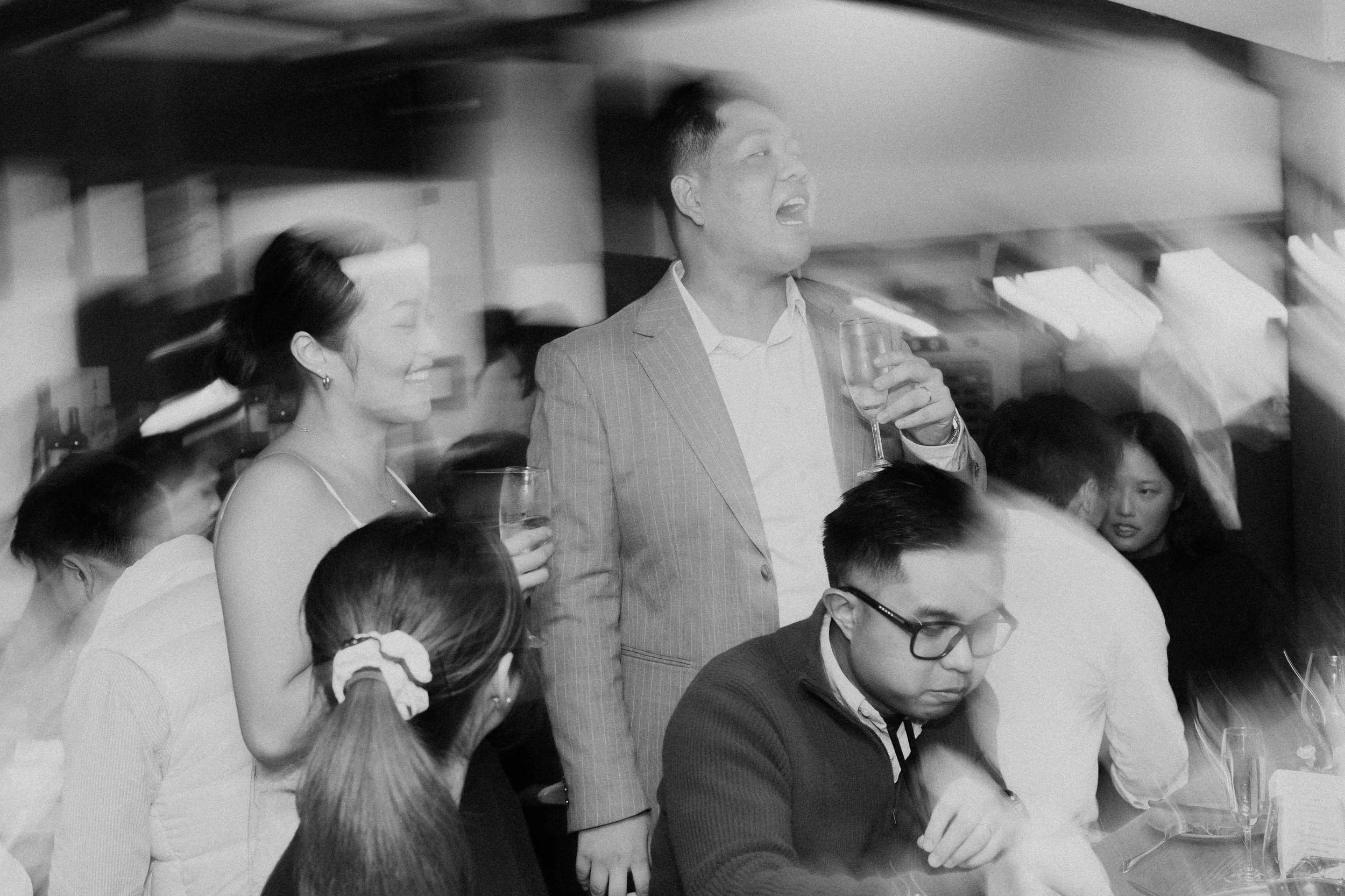 A black and white photo showing a group of people at a social gathering, with a man in a suit standing and holding a glass, smiling, while people around him are seated, engaged in conversation.