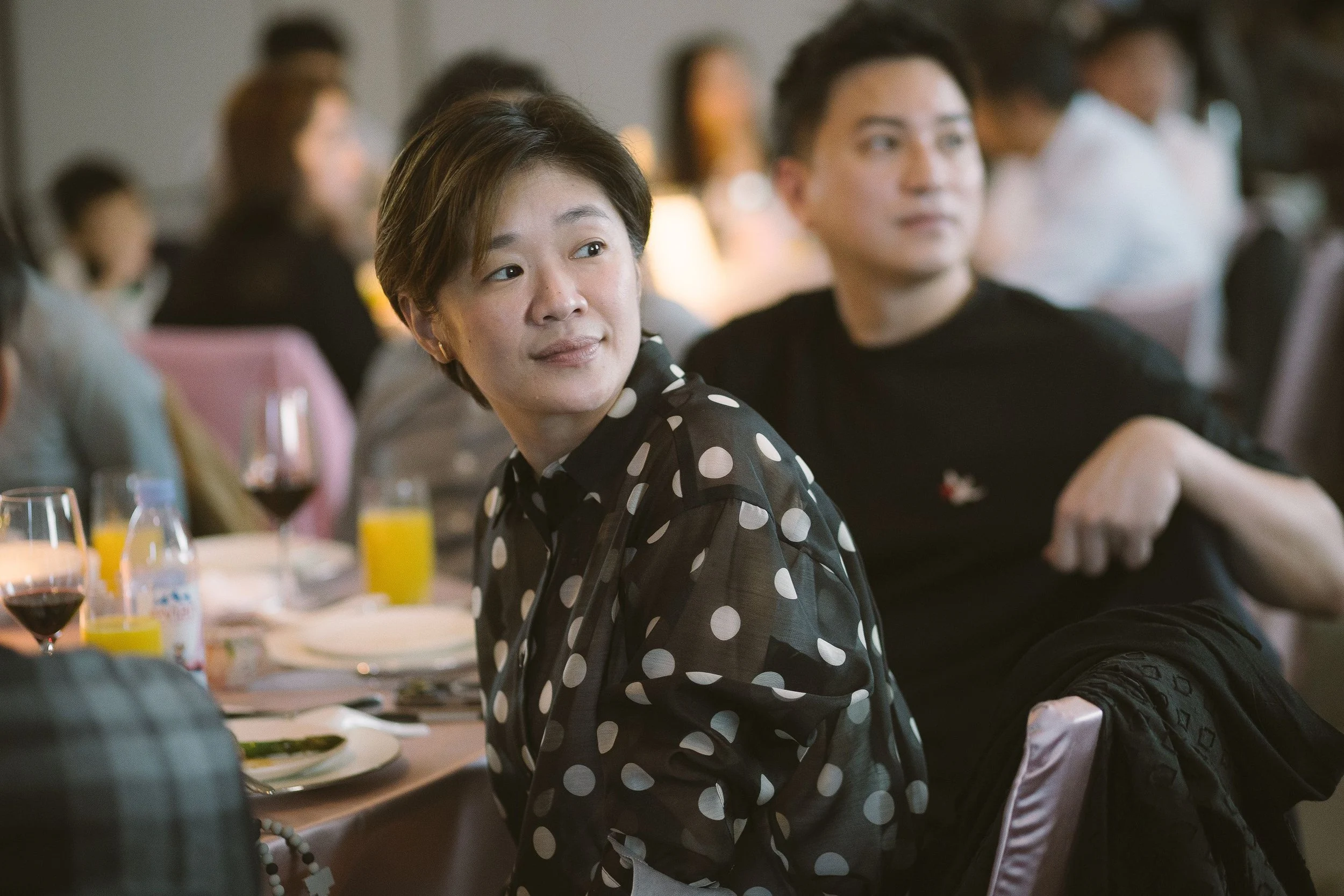 A woman with short brown hair wearing a black and white polka dot blouse sitting at a restaurant or banquet table during an event, with multiple people visible in the background.