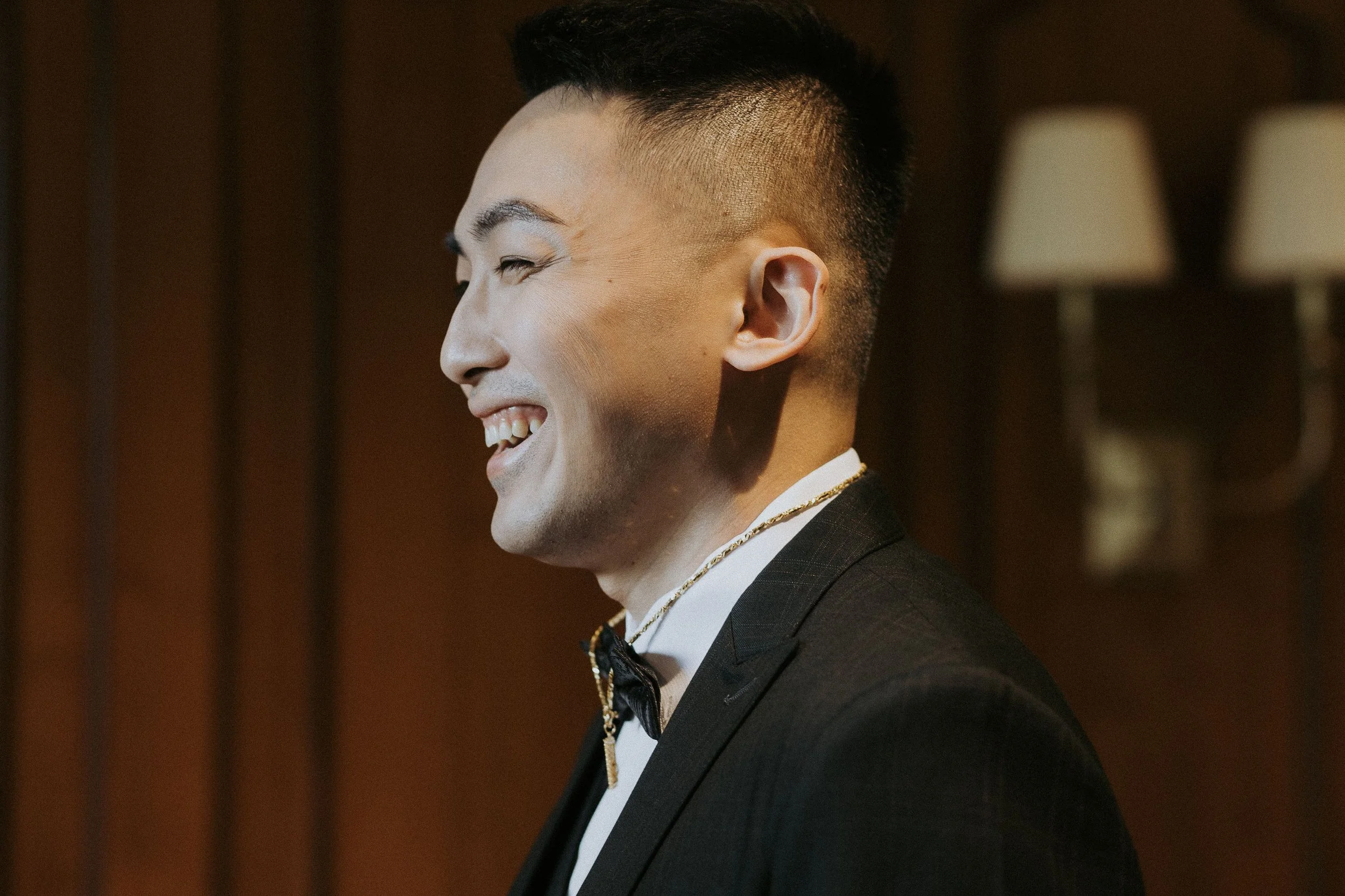 Side profile of a smiling man wearing a tuxedo with a gold necklace and bow tie, in a dimly lit indoor setting.