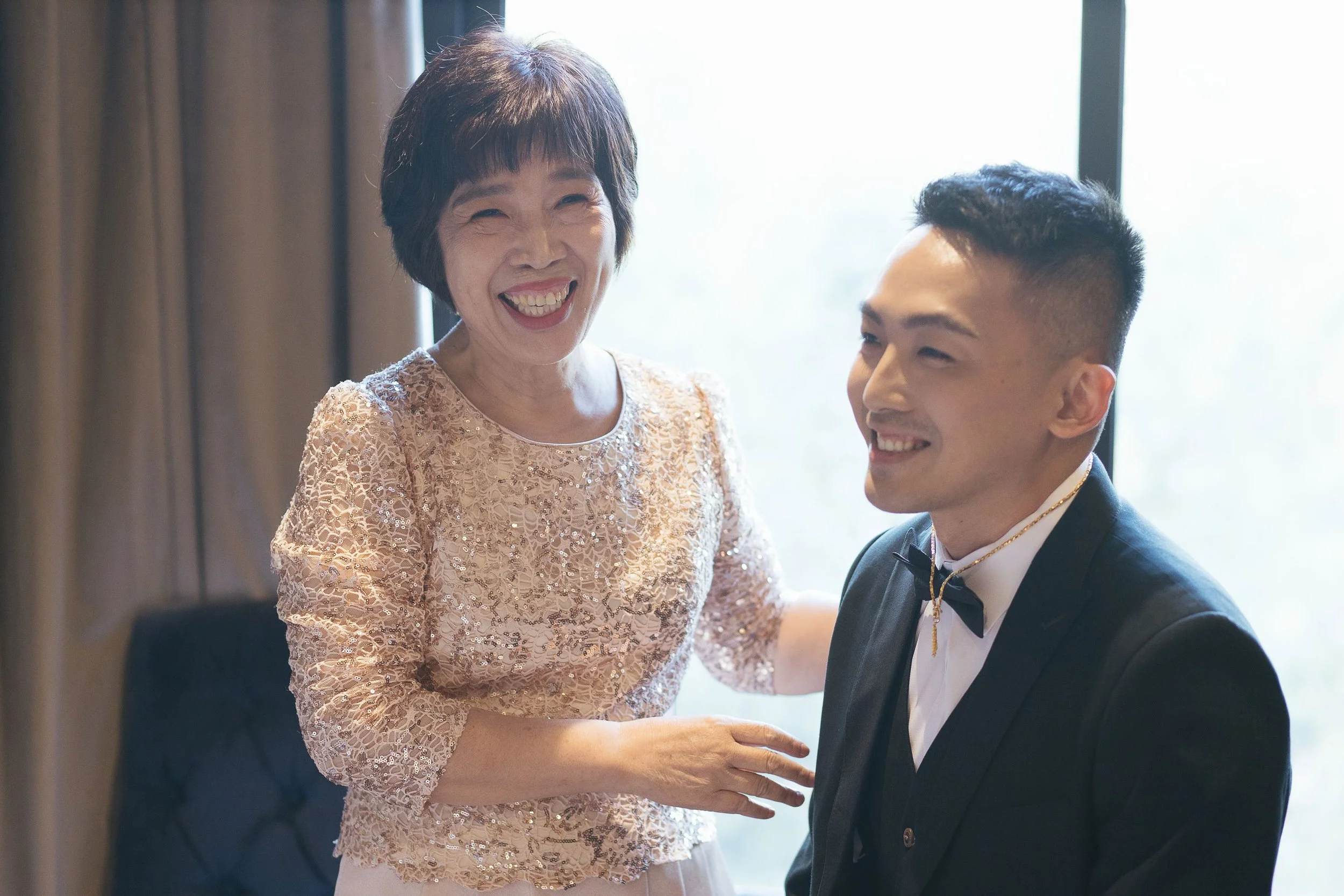 An older woman and a young man smiling and looking at each other. The woman has short dark hair and is wearing a peach lace top. The young man has short dark hair, is dressed in a tuxedo with a bow tie and a gold chain, and is sitting down. They appe