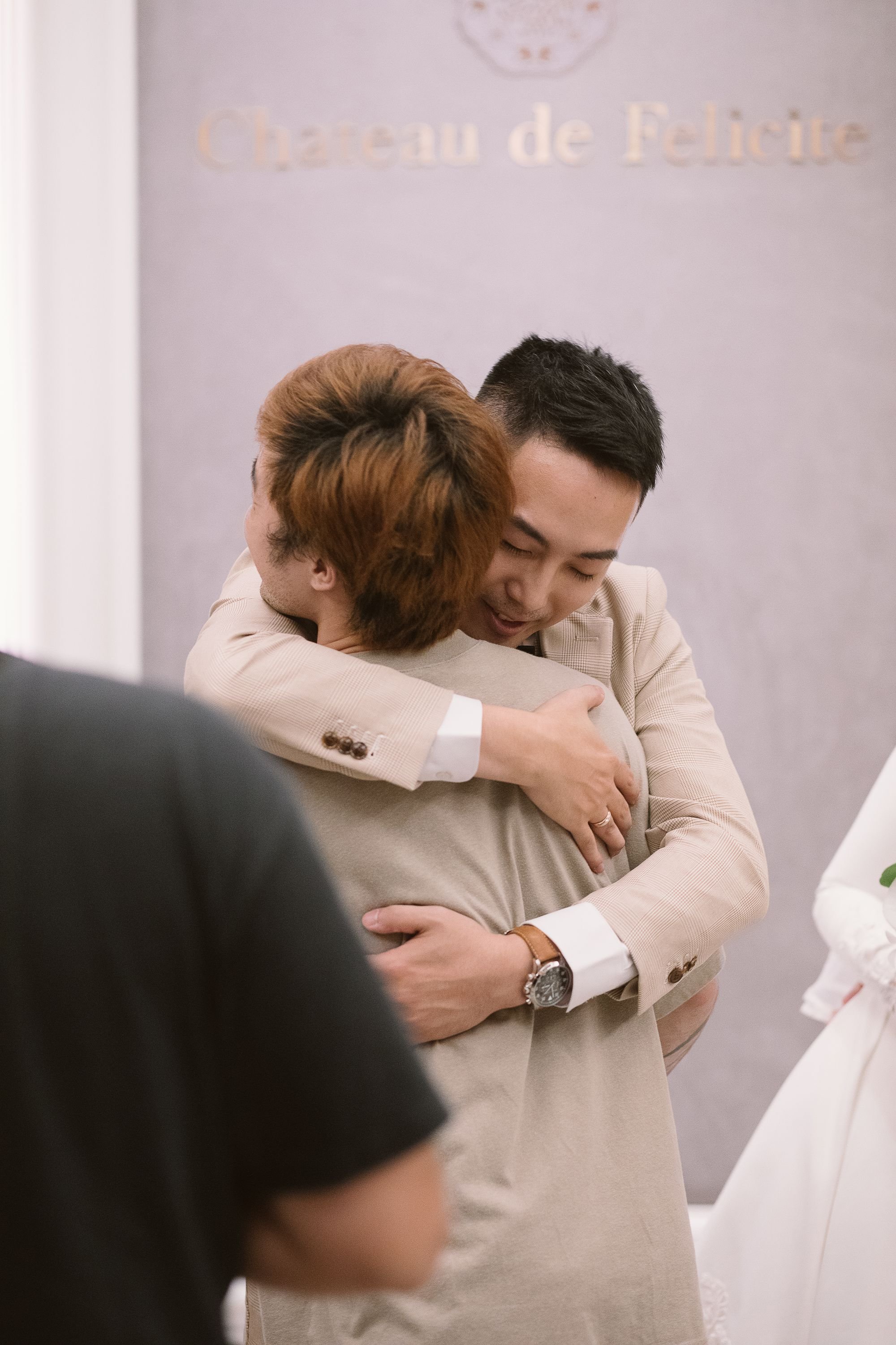 Two men are hugging each other warmly in a formal setting, with a blurred background that includes a sign reading 'Chateau de Felicite'.