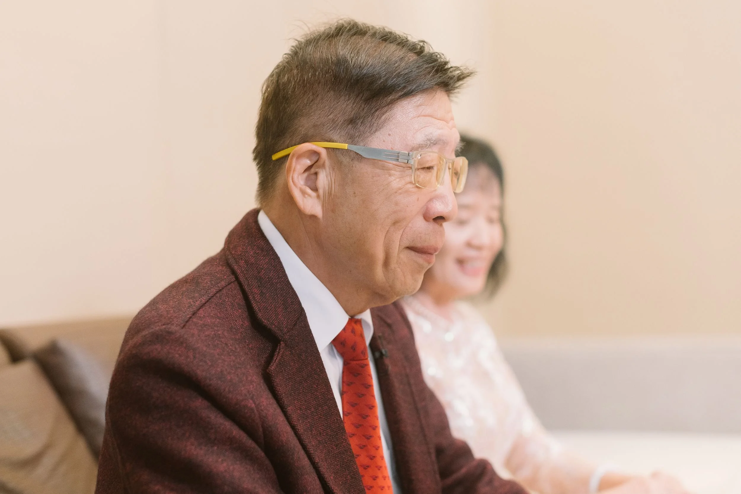 A man wearing glasses and a maroon suit with a red tie, sitting in a row with a woman in a white dress, both looking forward with closed eyes, possibly attending a formal event or ceremony.