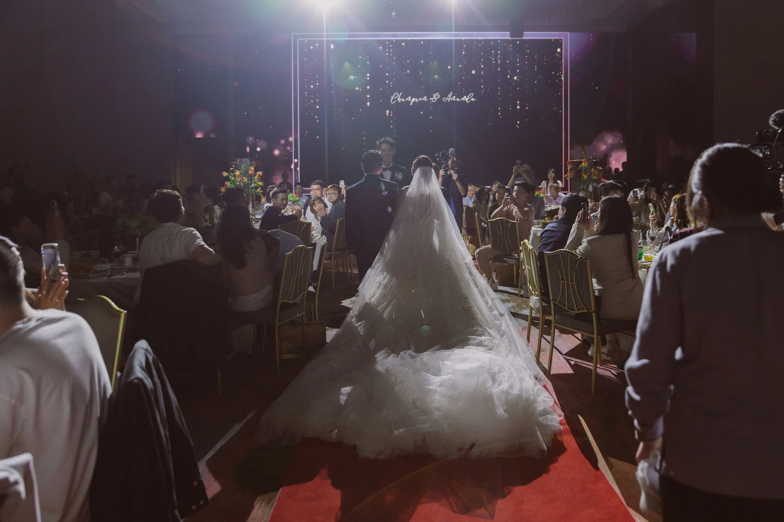 A wedding reception with a bride and groom walking down the aisle, surrounded by guests seated at tables. The room is dimly lit with a large digital display in the background showing a starry sky and the names 'Champun & Ahad'. The bride wears a whit