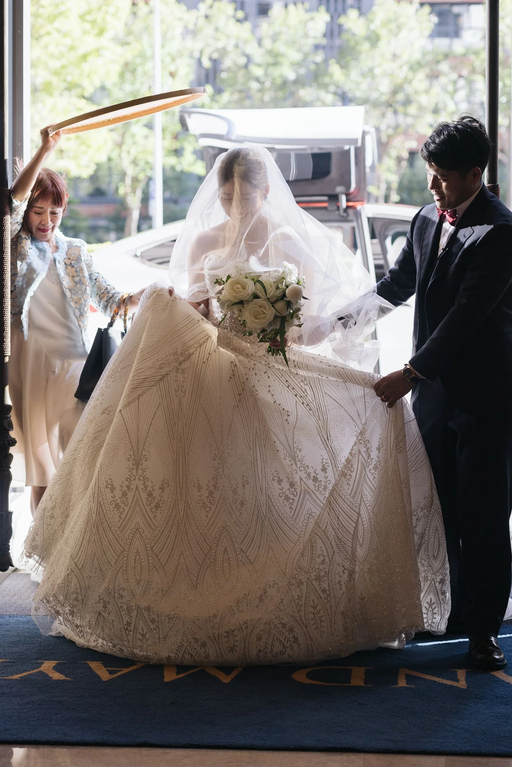 Bride in a wedding gown with veil holding a bouquet of white roses, standing with groom dressed in a black tuxedo, while a woman in a light-colored dress and floral jacket adjusts her train at the entrance of a building with glass doors.