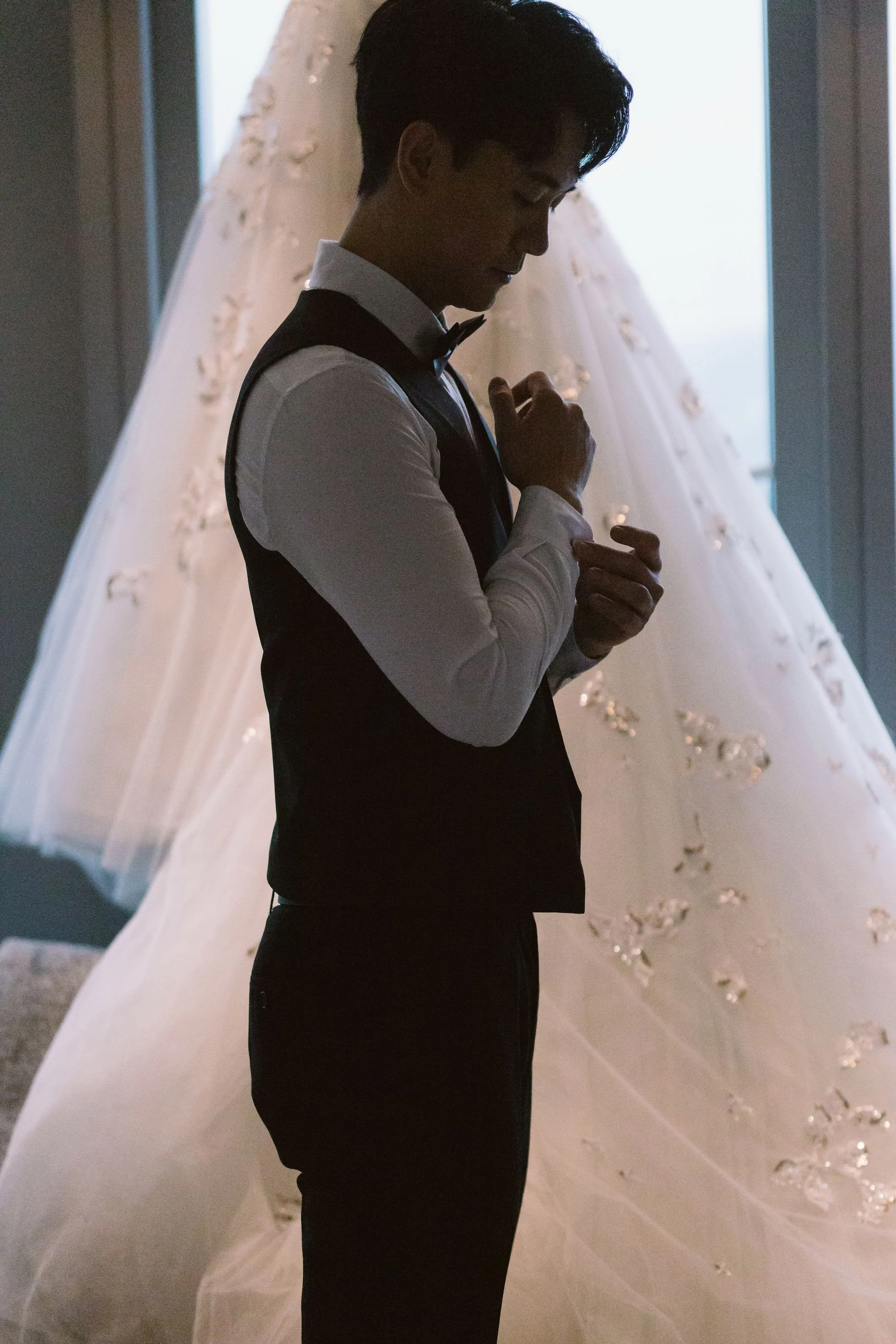A young man poised with his head bowed and eyes closed, adjusting his bow tie in front of a large wedding dress with floral details.