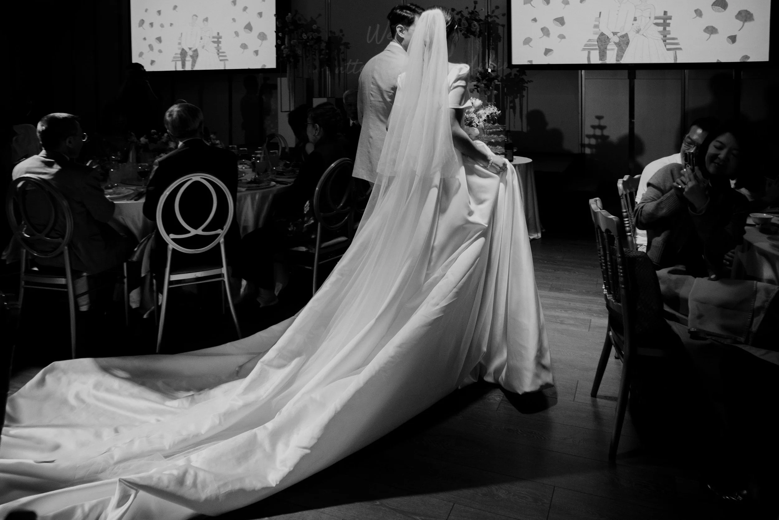 Black and white photo of a bride in a wedding gown and veil, sitting on a chair during a wedding reception. Guests are seated at tables around her, and a large screen in the background displays an illustration of a couple on a bench surrounded by lea