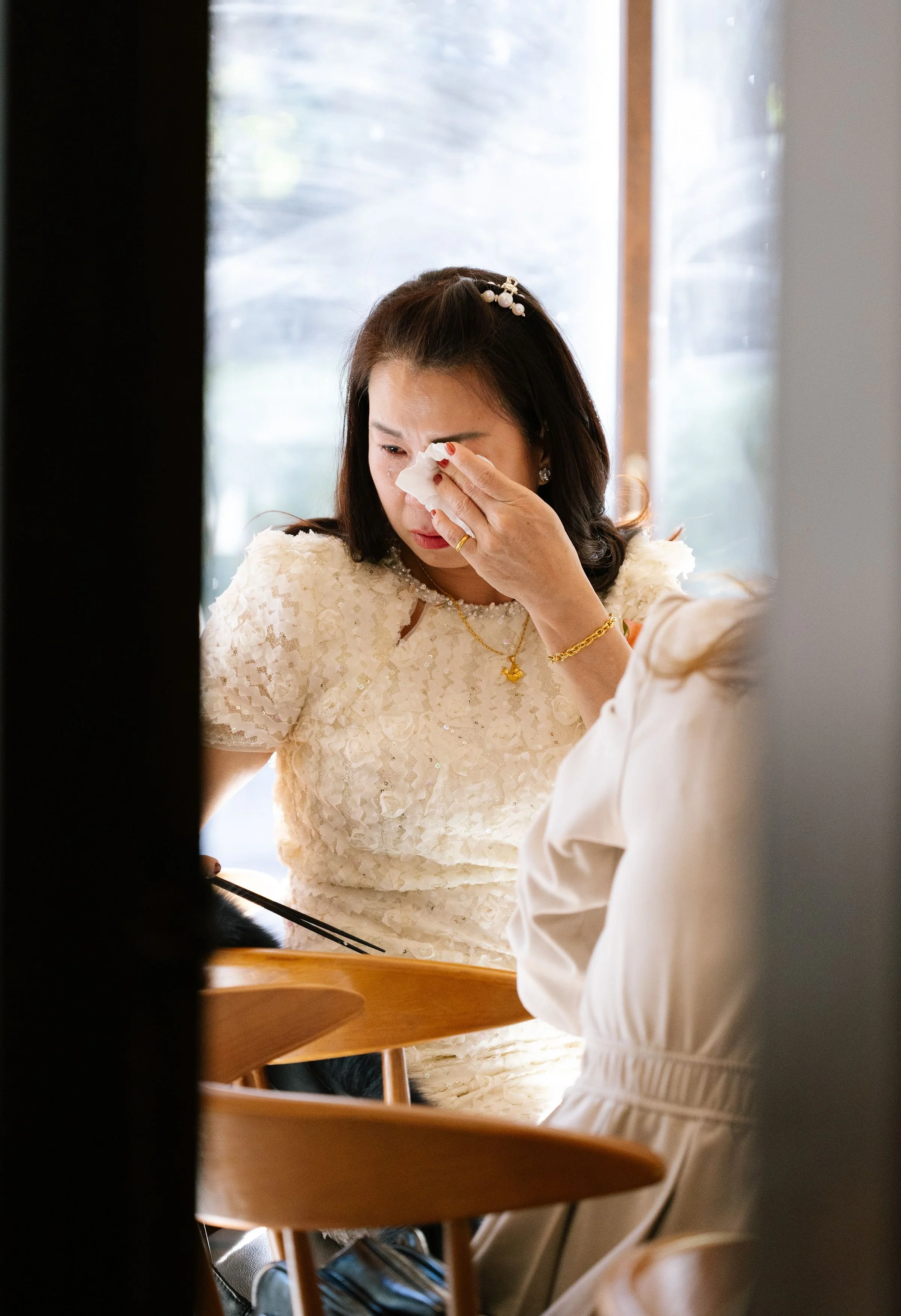 A woman in a cream-colored lace dress is wiping tears from her eyes with a tissue. She is seated at a wooden table, wearing jewelry and a hairpin, in front of a window with sunlight.