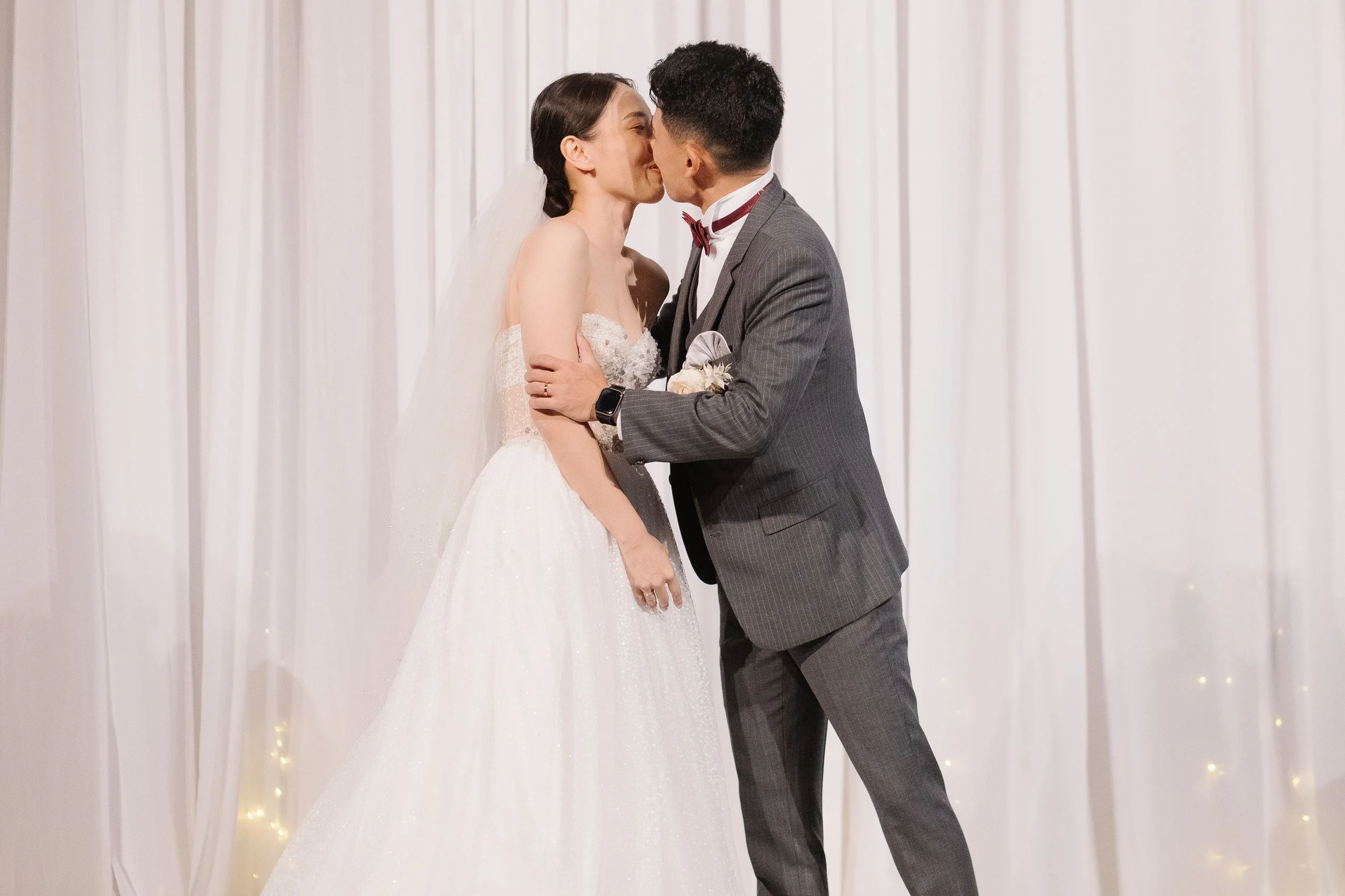A bride and groom share a kiss during their wedding ceremony in front of a white curtain backdrop.