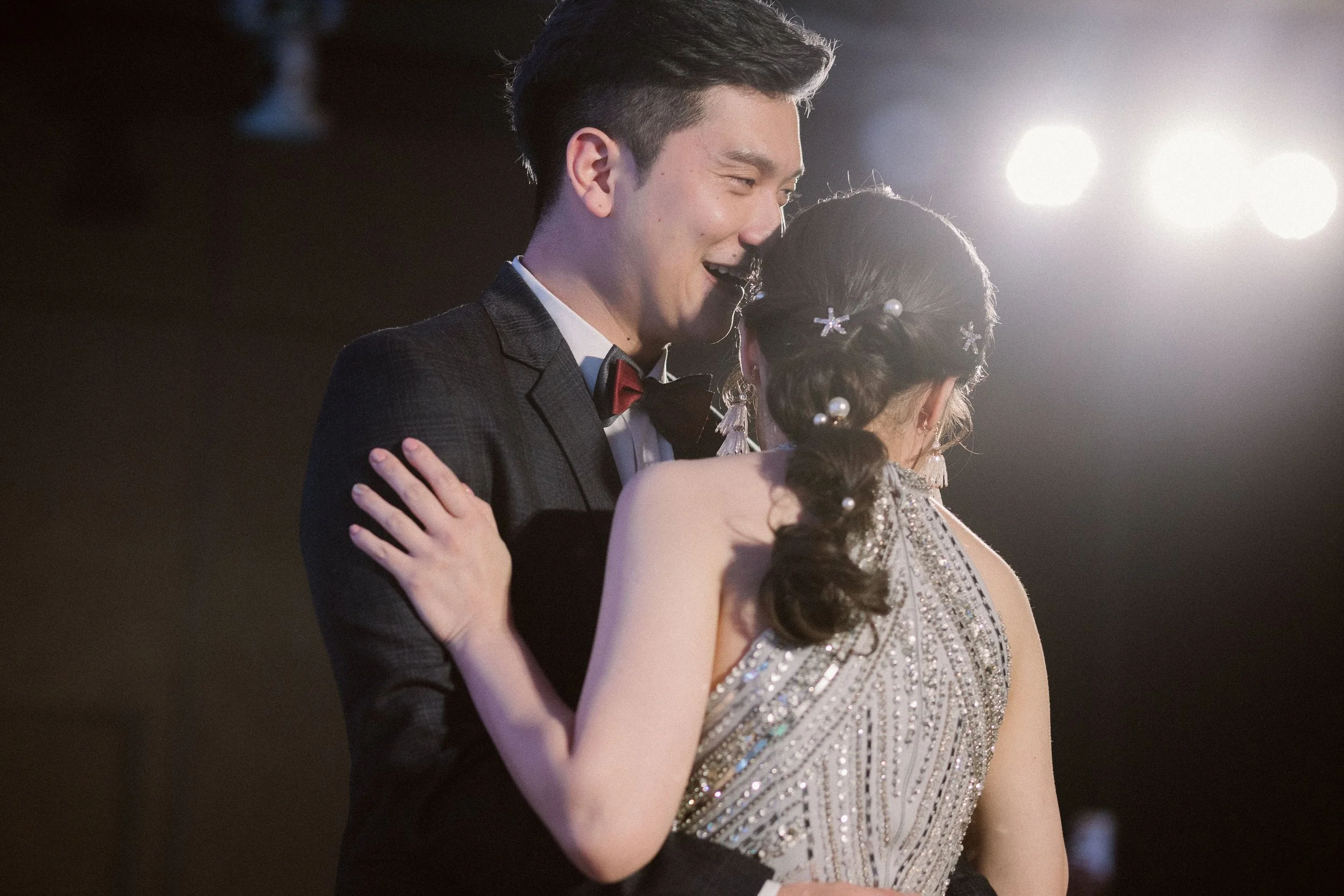 A young man in a tuxedo with a red bow tie dancing closely with a young woman in a bead-embellished dress at a formal event, both smiling and embracing each other.