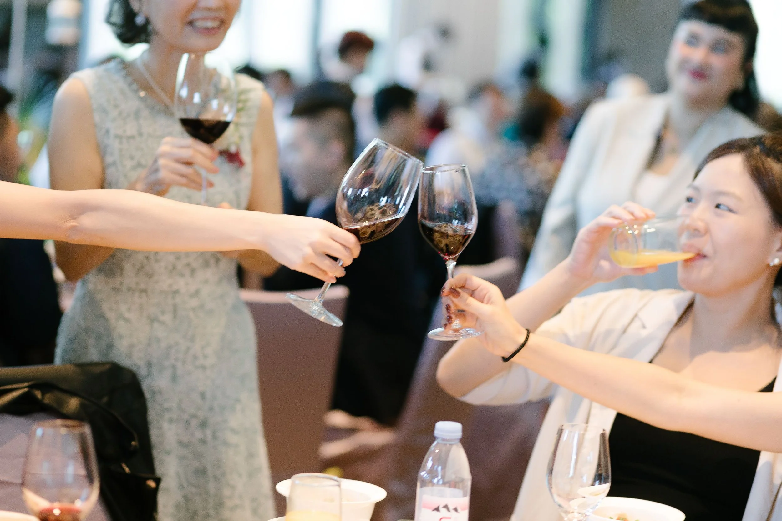 People at a social gathering holding glasses of red wine and drinks, with one woman in a sleeveless dress smiling and pouring wine.