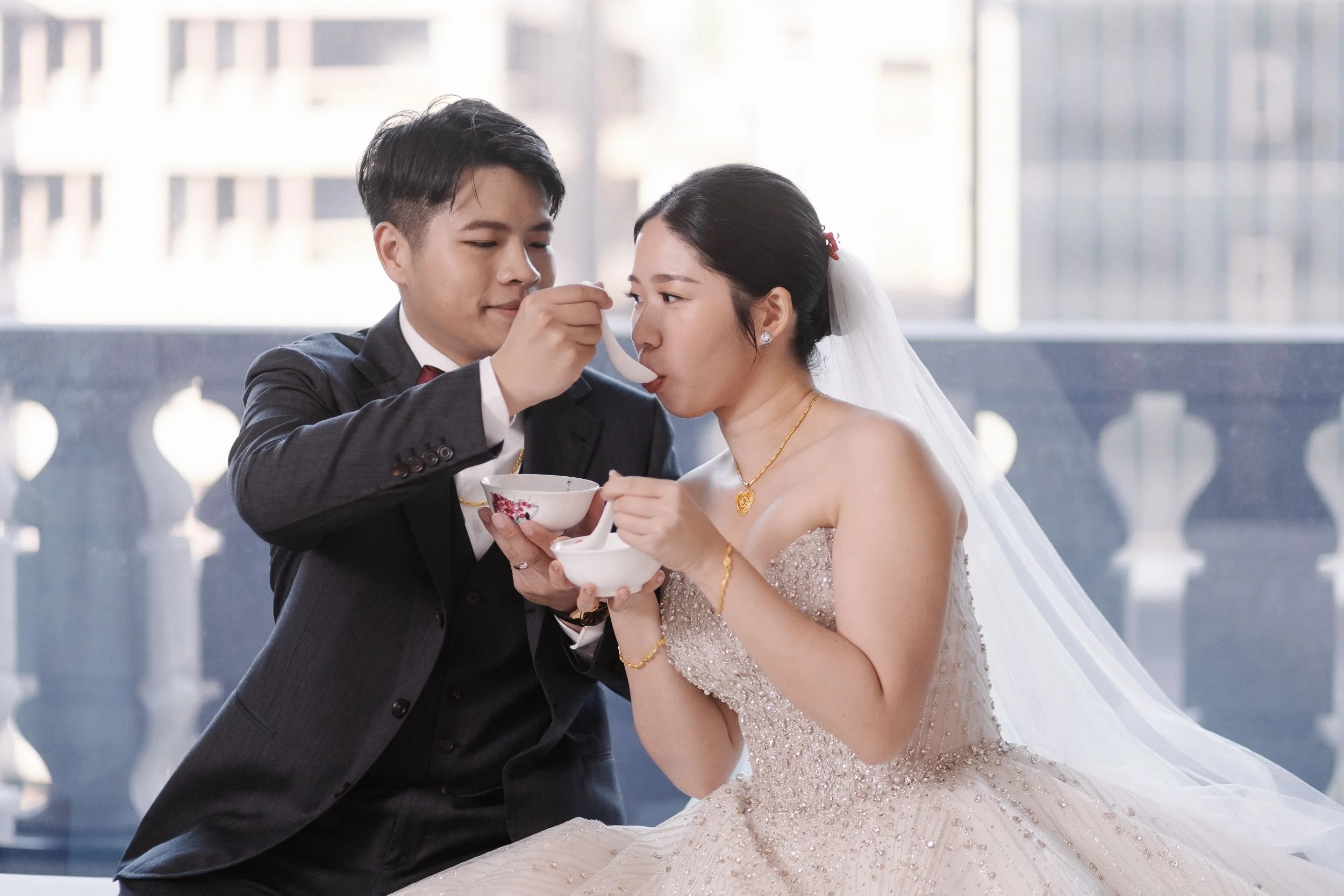 A groom helping a bride eat wedding cake at their wedding reception, with a cityscape background visible through large windows.
