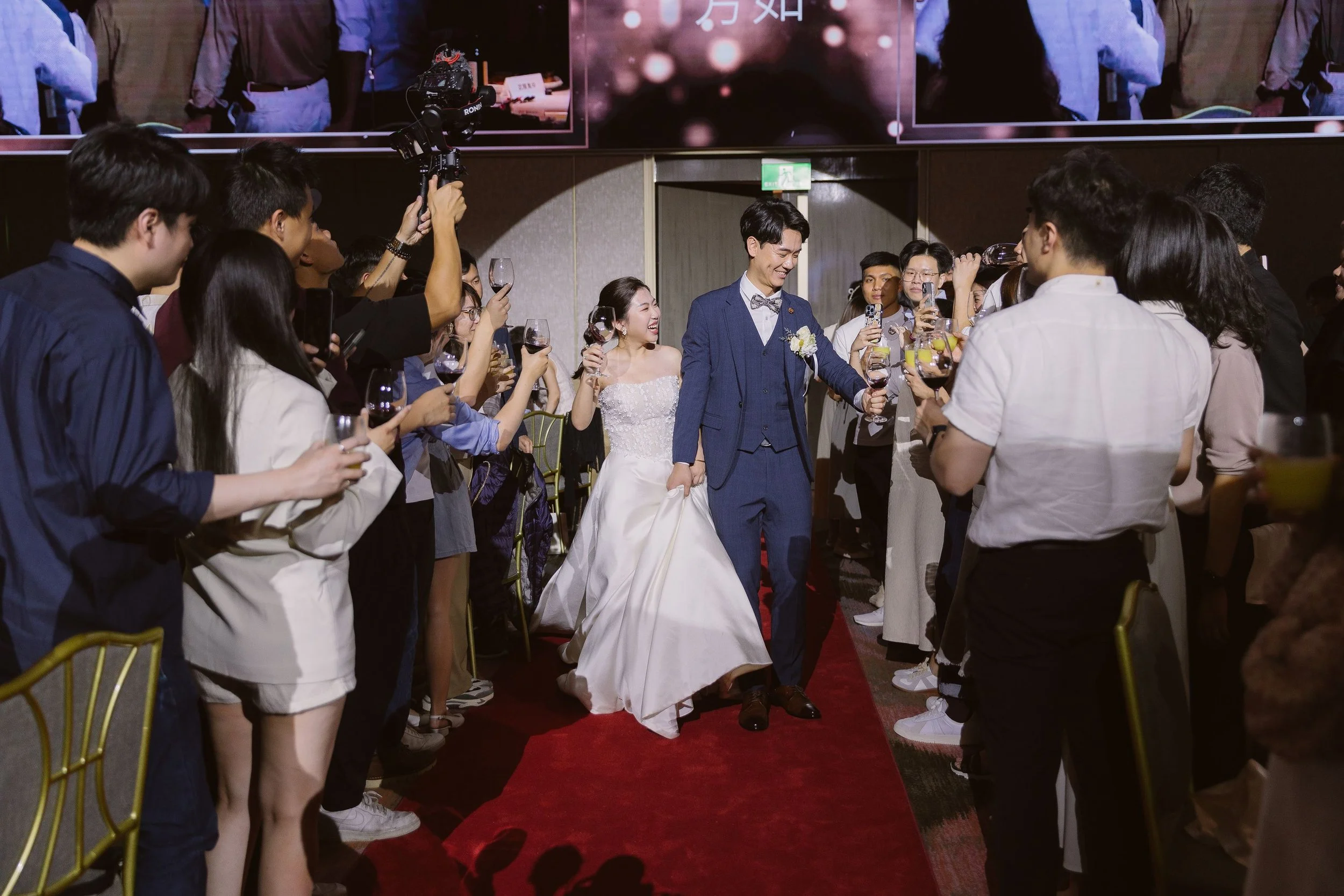 A bride and groom walking down a red aisle, surrounded by guests holding glasses of wine and taking photos at their wedding reception.