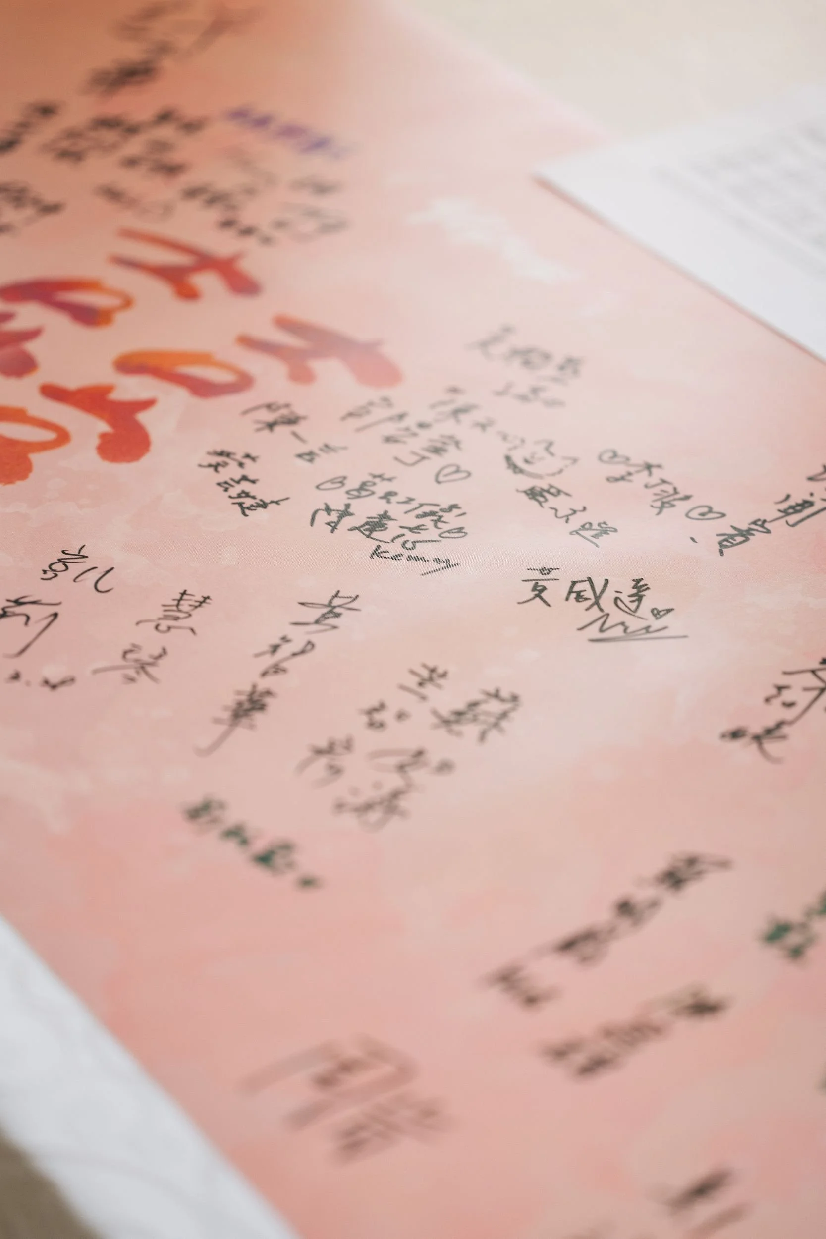 Close-up of handwritten messages and signatures on pink paper, with some Japanese characters visible and a blurred white paper in the background.