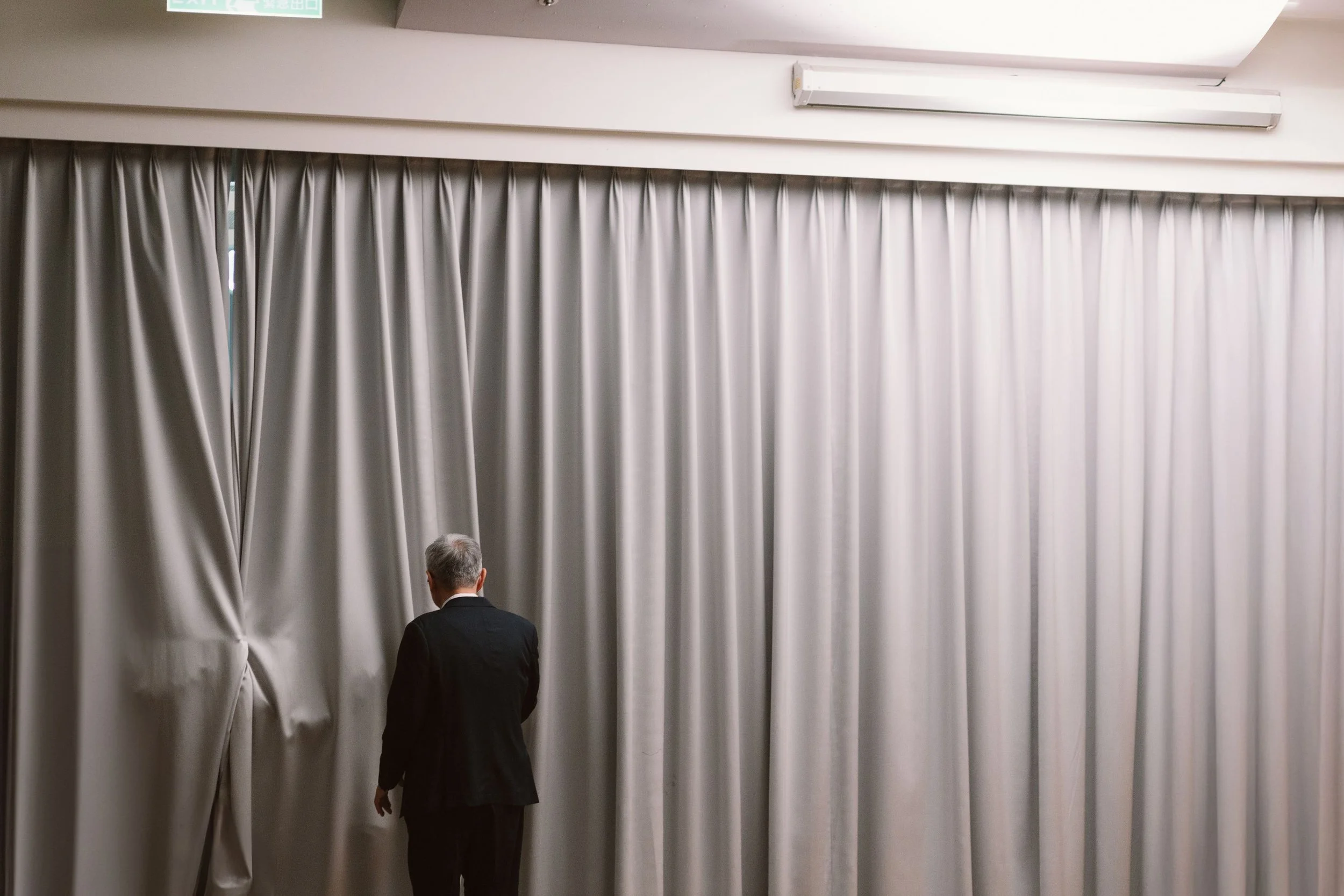 A man in a black suit standing in front of closed gray curtains in an indoor setting, with part of an emergency exit sign visible at the top.